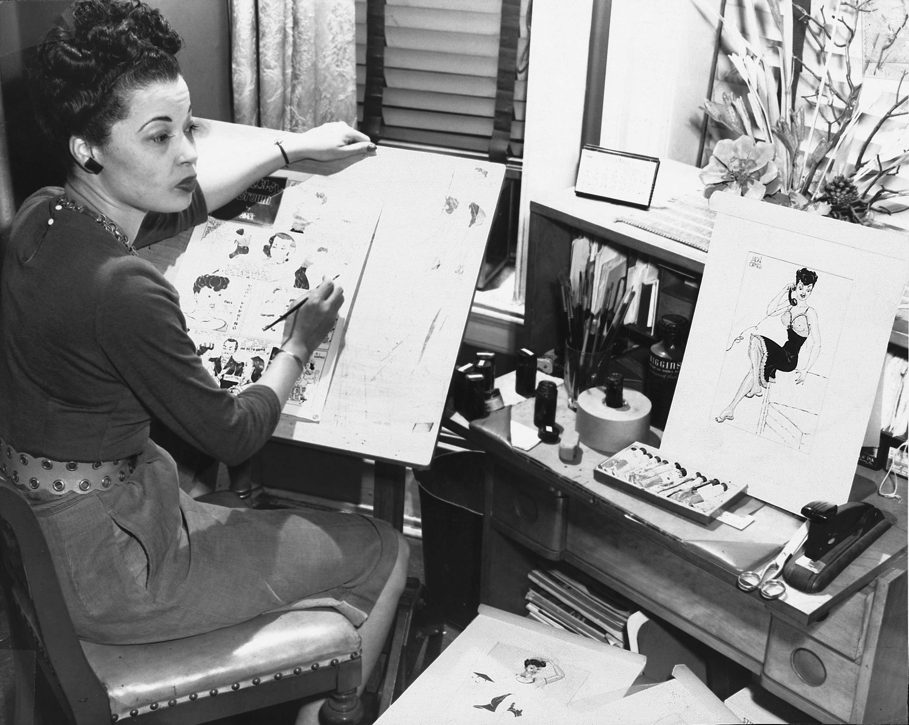 A black and whtie photo of Jackie Ormes at working. She is drawing on one paper while looking at inspiration.