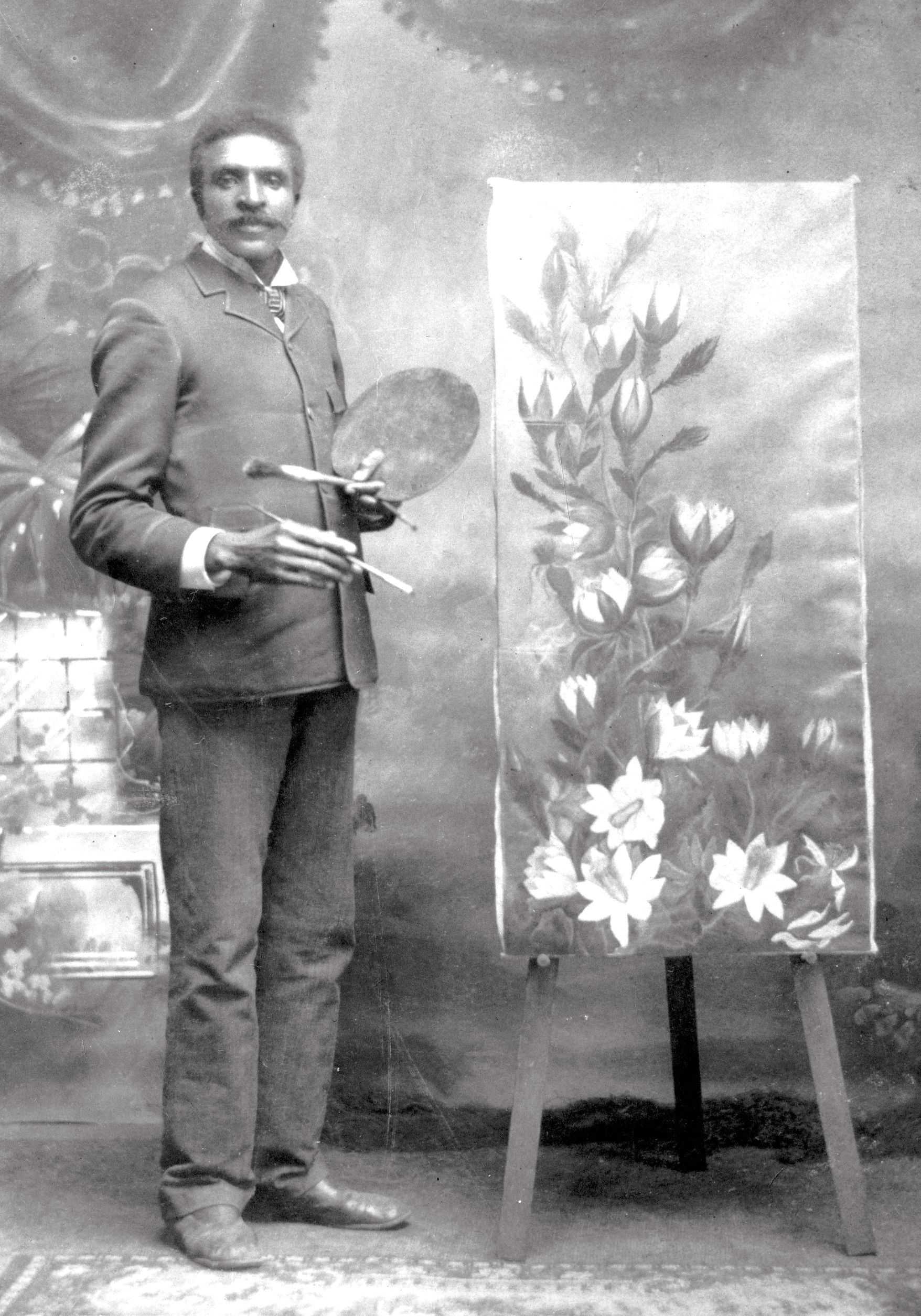 Black and white photograph of a young George Washington Carver holding an artists palette, standing next to a floral painting.