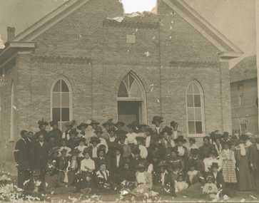 A black-and-white photographic postcard of men, women, and children gathered in front of a church. There are losses at TC and TL.