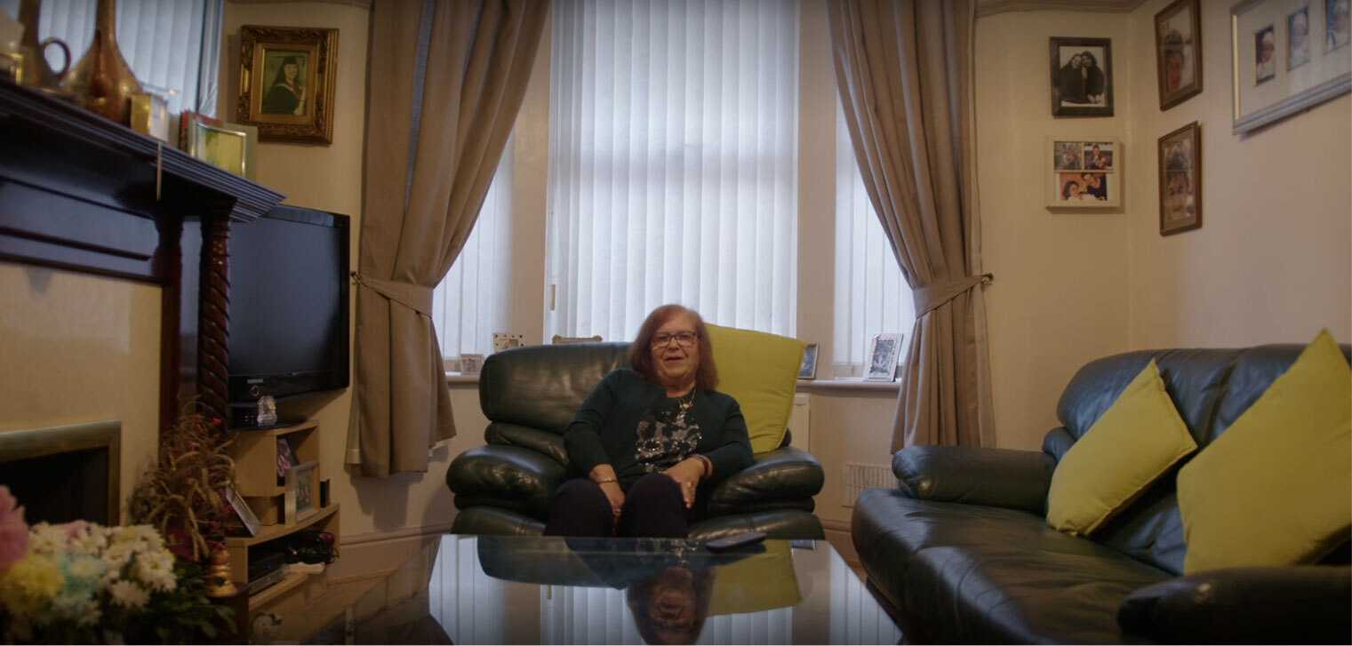 Person seated in a leather armchair in a living room with large curtained windows, framed family photos on the walls, and matching sofas on either side.
