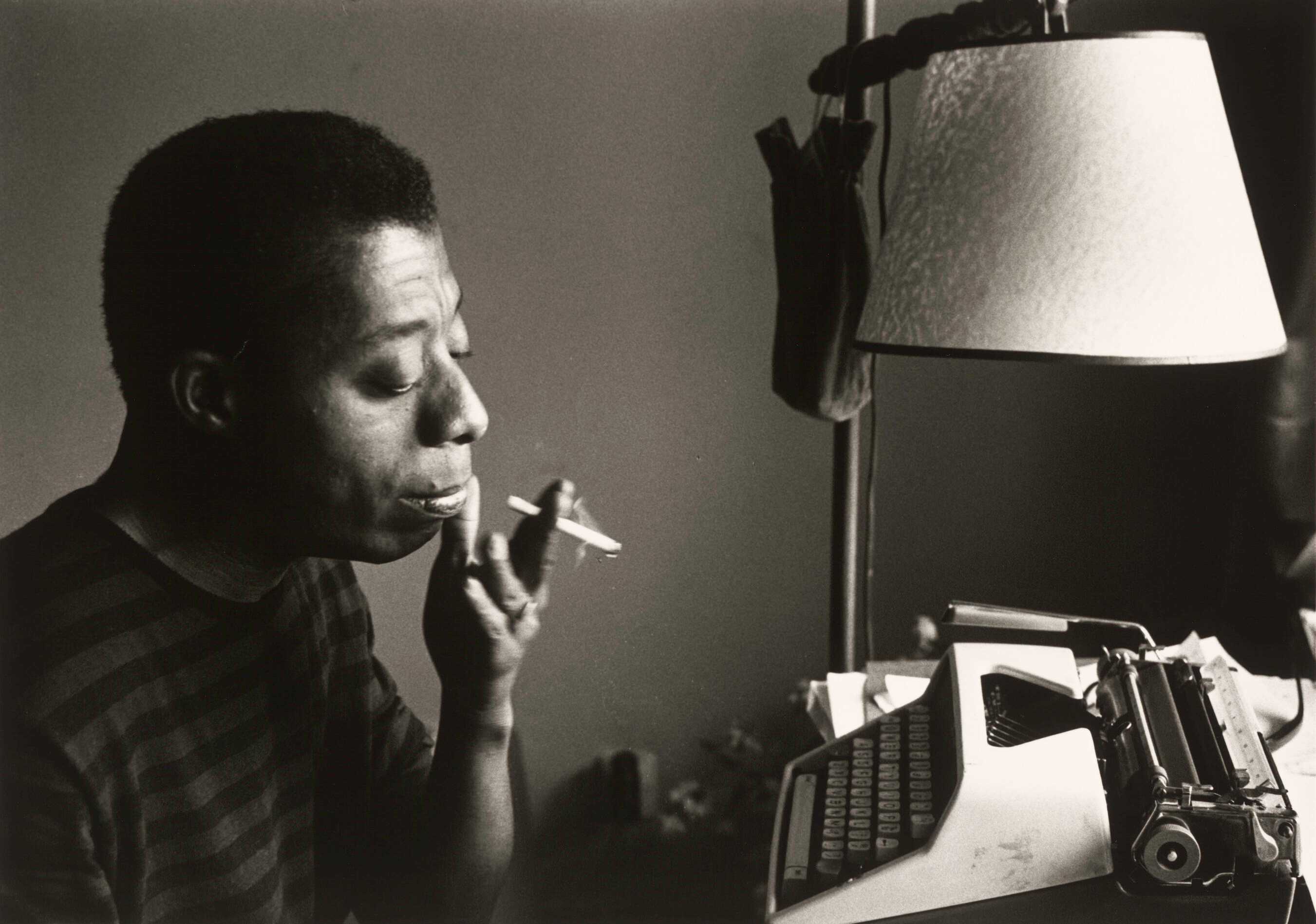 A photograph of James Baldwin sitting at a typewriter and smoking a cigarette.