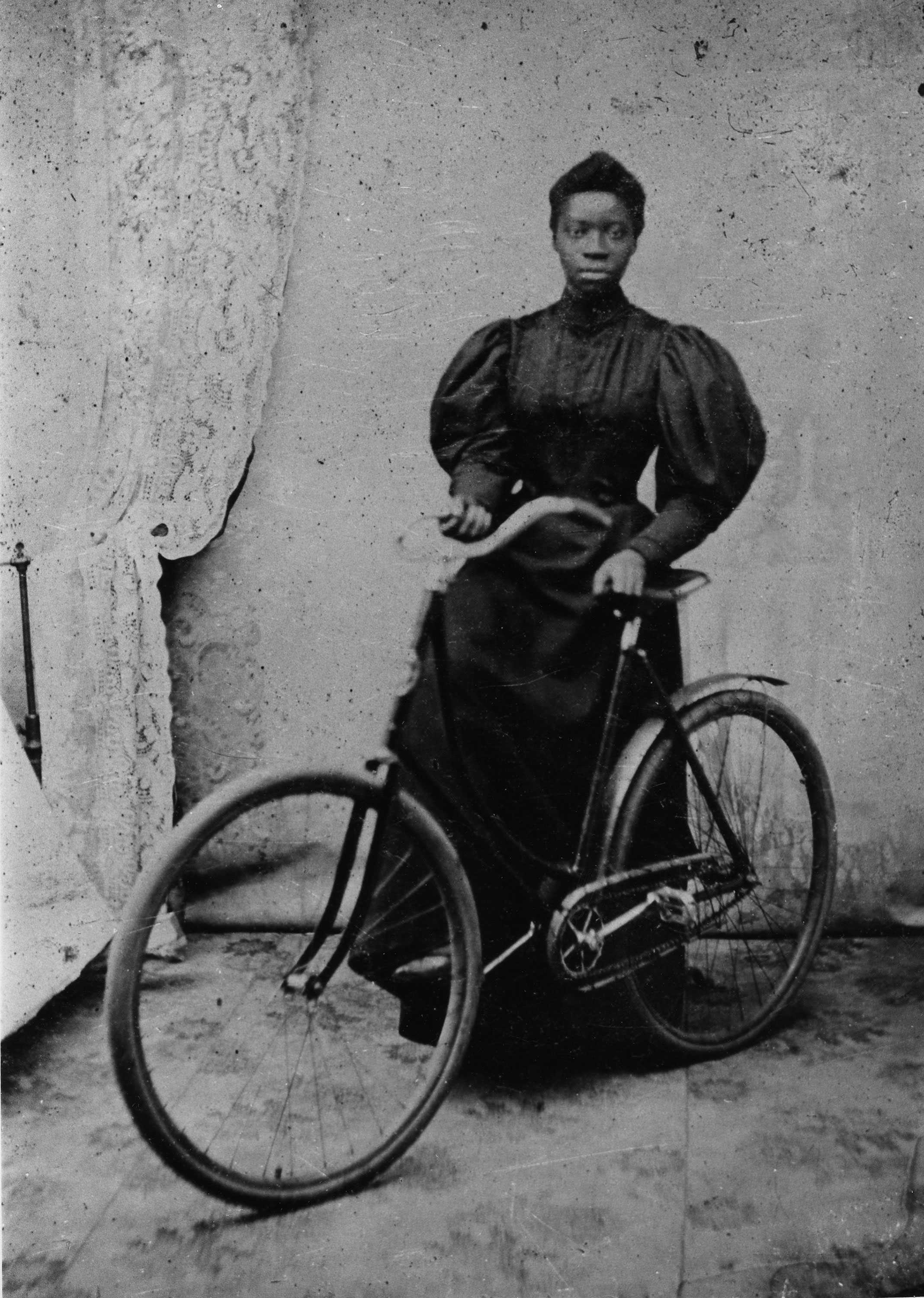 A black and white photo of a woman standing with her bicycle. She is slightly out of focus