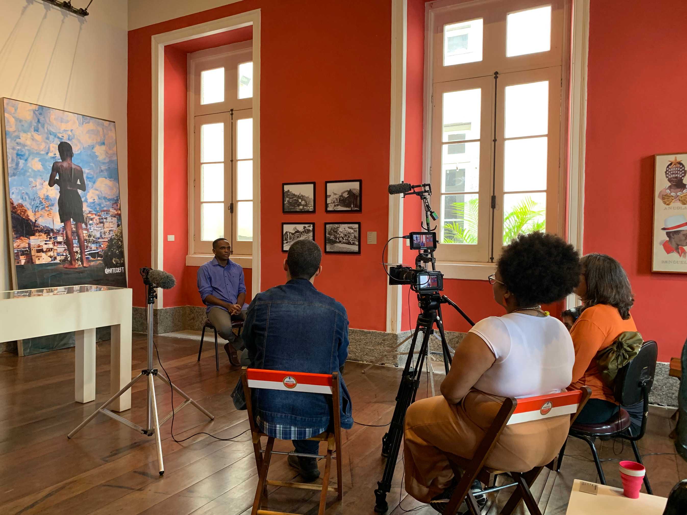 A person sits for a filmed interview in an art gallery with red walls, while a small audience, camera, and microphones record the discussion beside large artworks and tall windows.