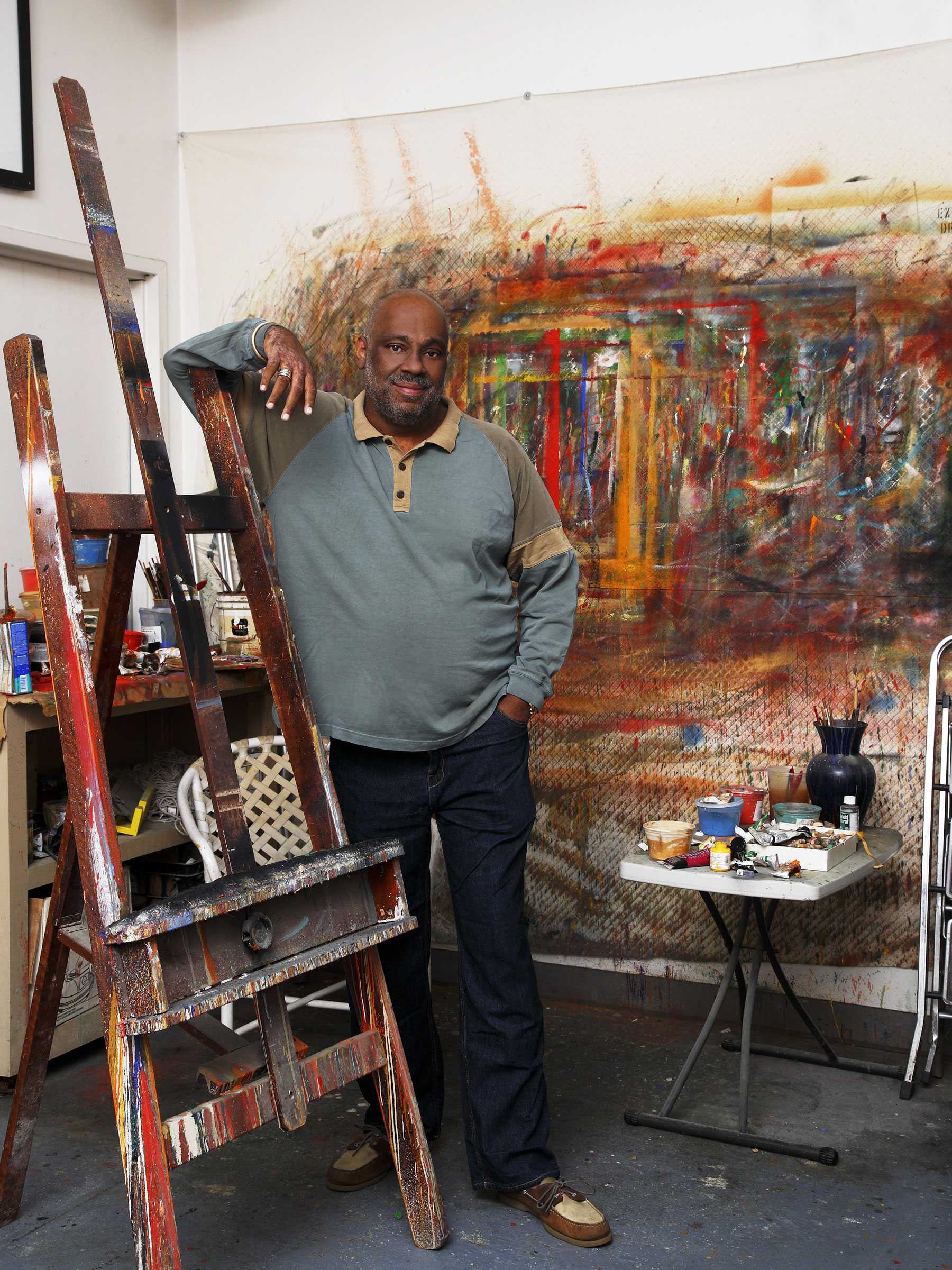 Man standing in an artist's studio, leaning on a paint-splattered easel with a colorful abstract painting in the background.