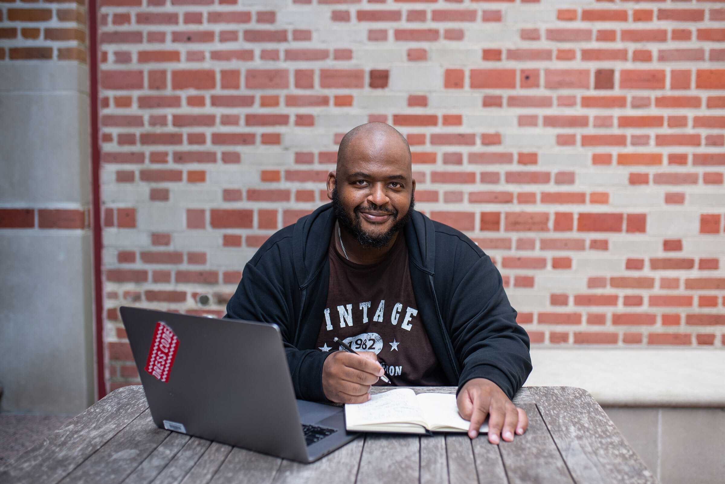 Color photograph of writer Kiese Laymon seated at a table with an open book and computer.