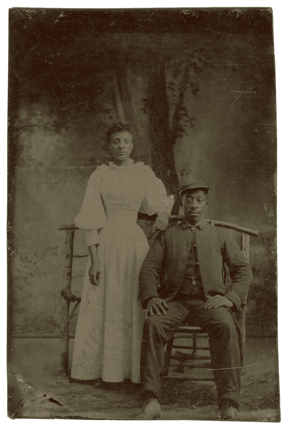 Tintype photograph depicting a seated soldier with woman standing next to him. Woman is wearing long white dress with puffy sleeves.