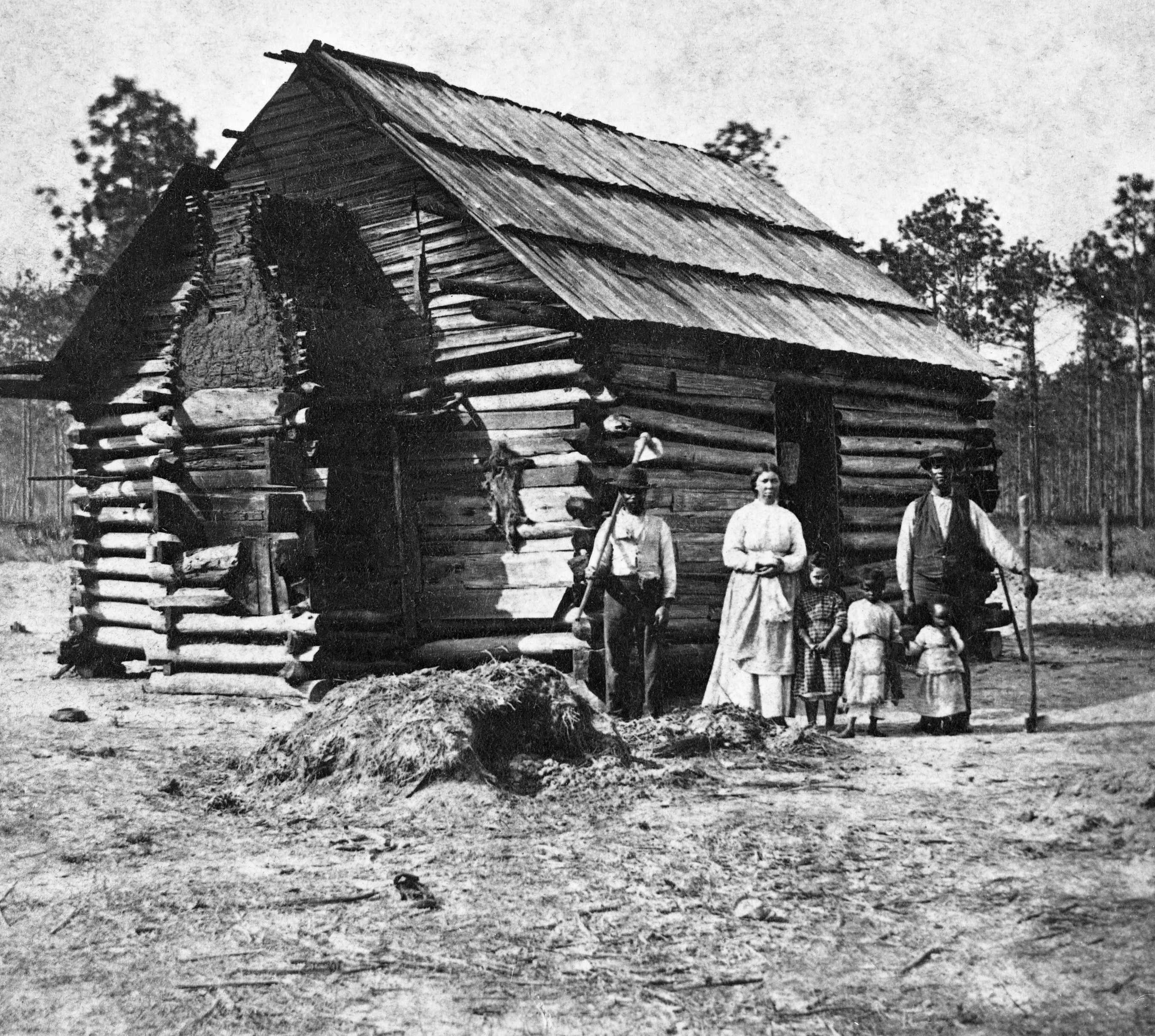A black and white photo of a family of 5 posing for a photo in front a small log cabin.