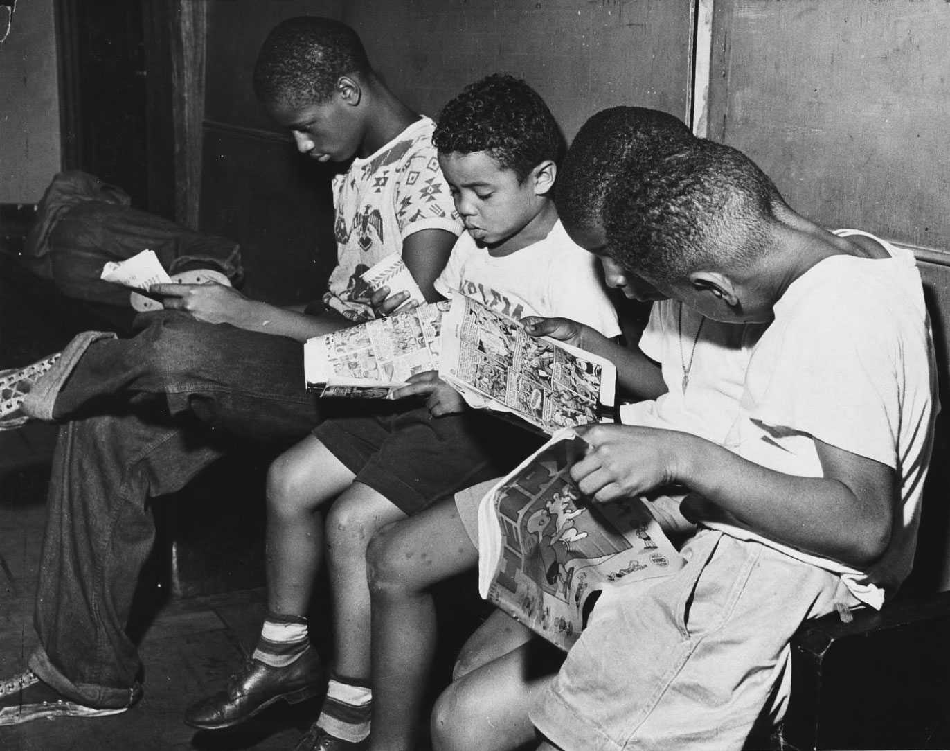 A black and white photo of 4 yound black boys sitting together, each reading a comic book.