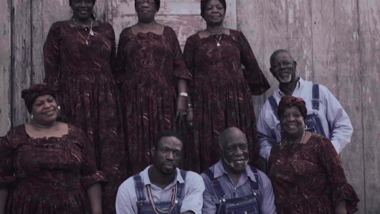 A group of McIntosh County Shouters pose together in costume in front of a wood panel structure.