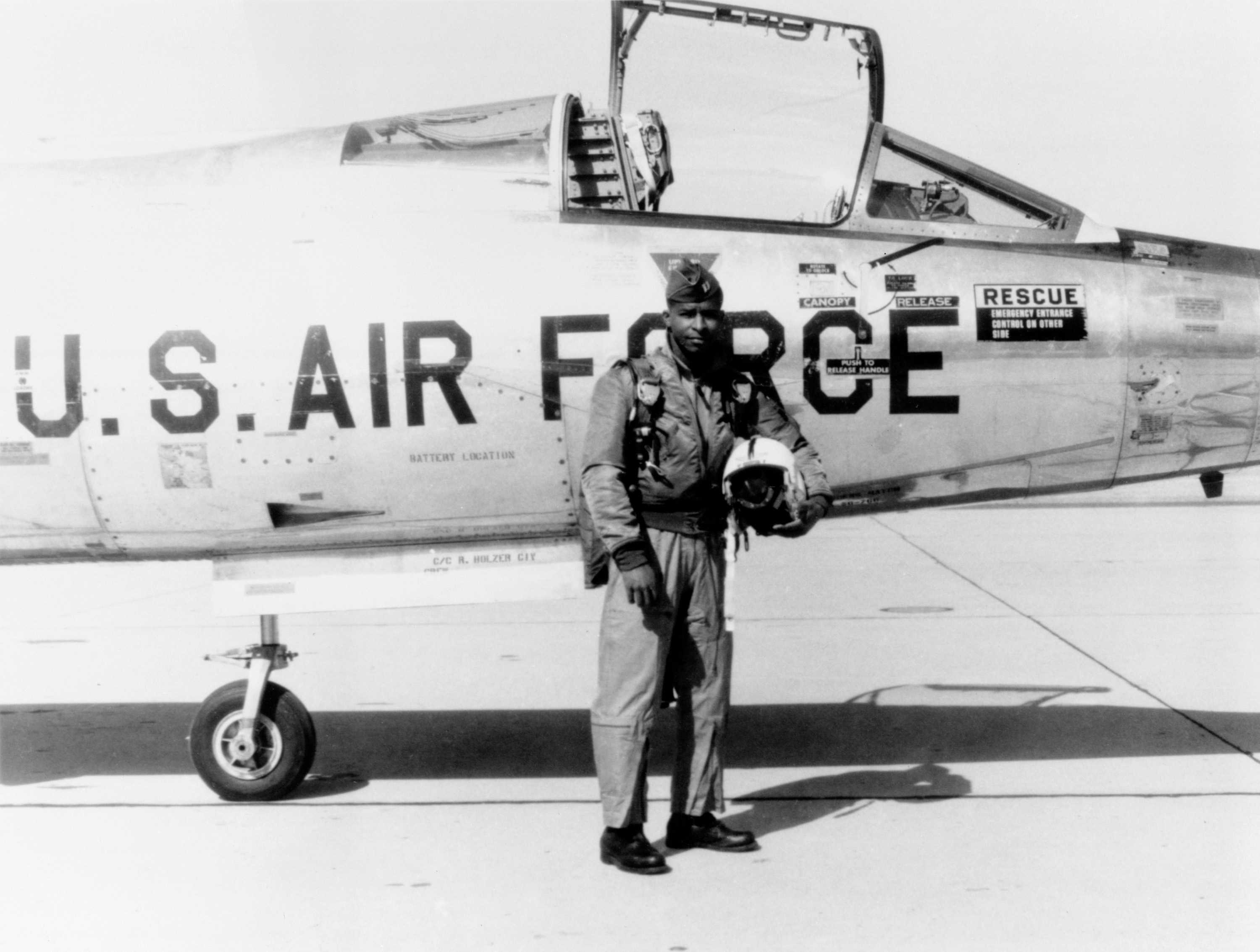 Lawrene poses for a black and white photo in front of a U.S Air Force plane. He is dressed in his flying suit.