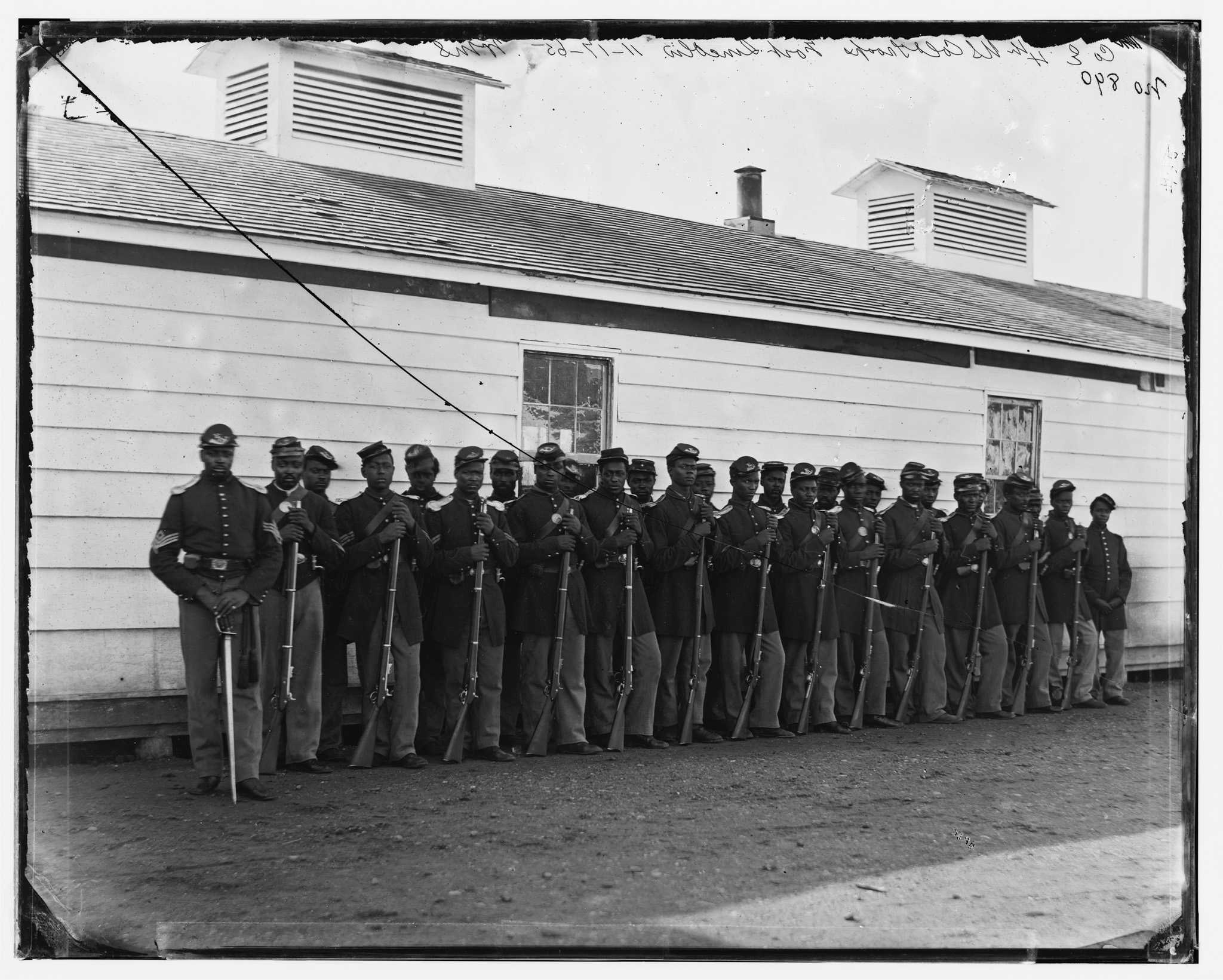 Black and white photo of the Company E, 4th U.S. Colored Infantry standing before a building.