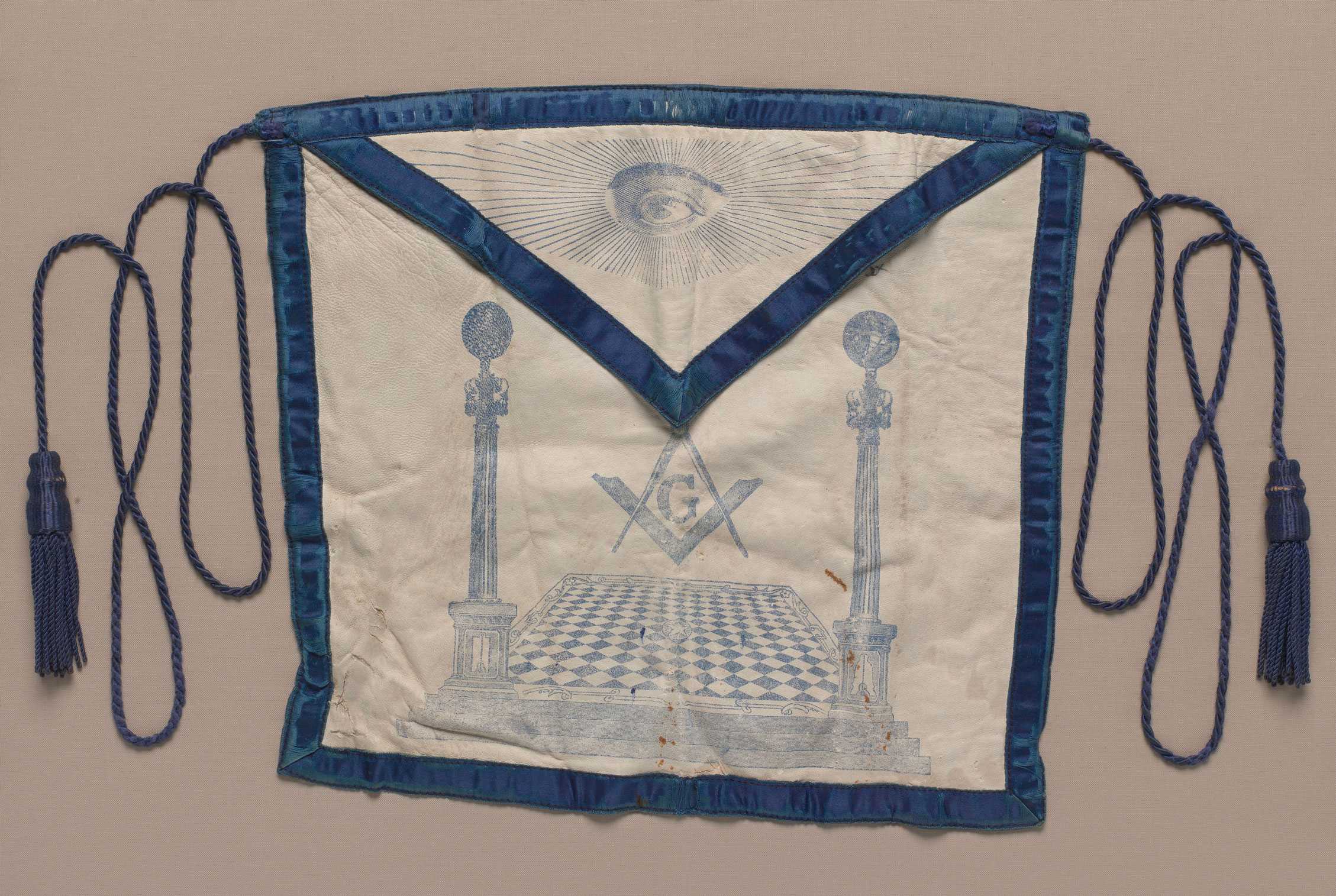 A blue trimmed Masonic apron with two tassels and a print of an eye and open hall.