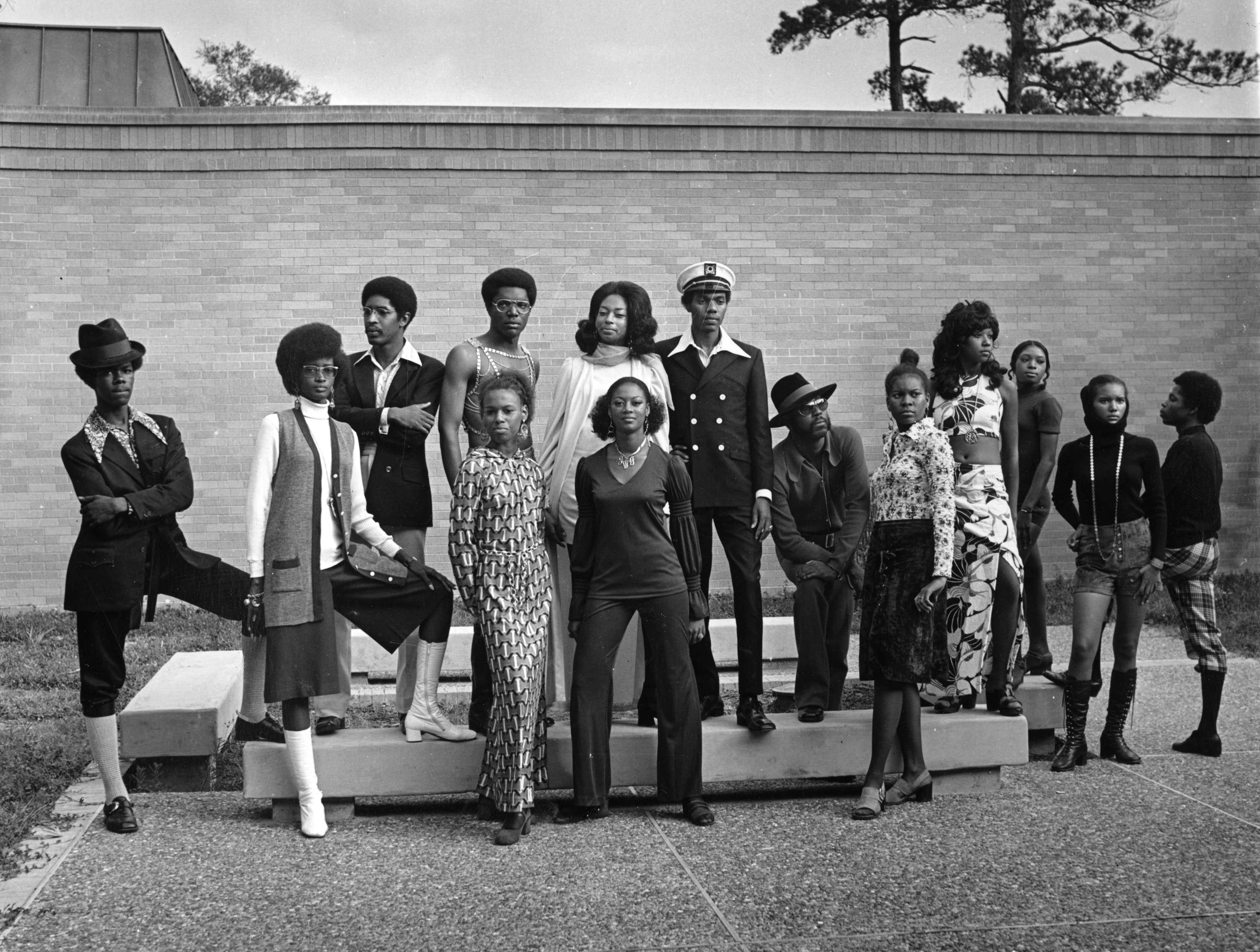 Black-and-white photo of a group of people in 1970s fashion posing on steps against a brick wall.