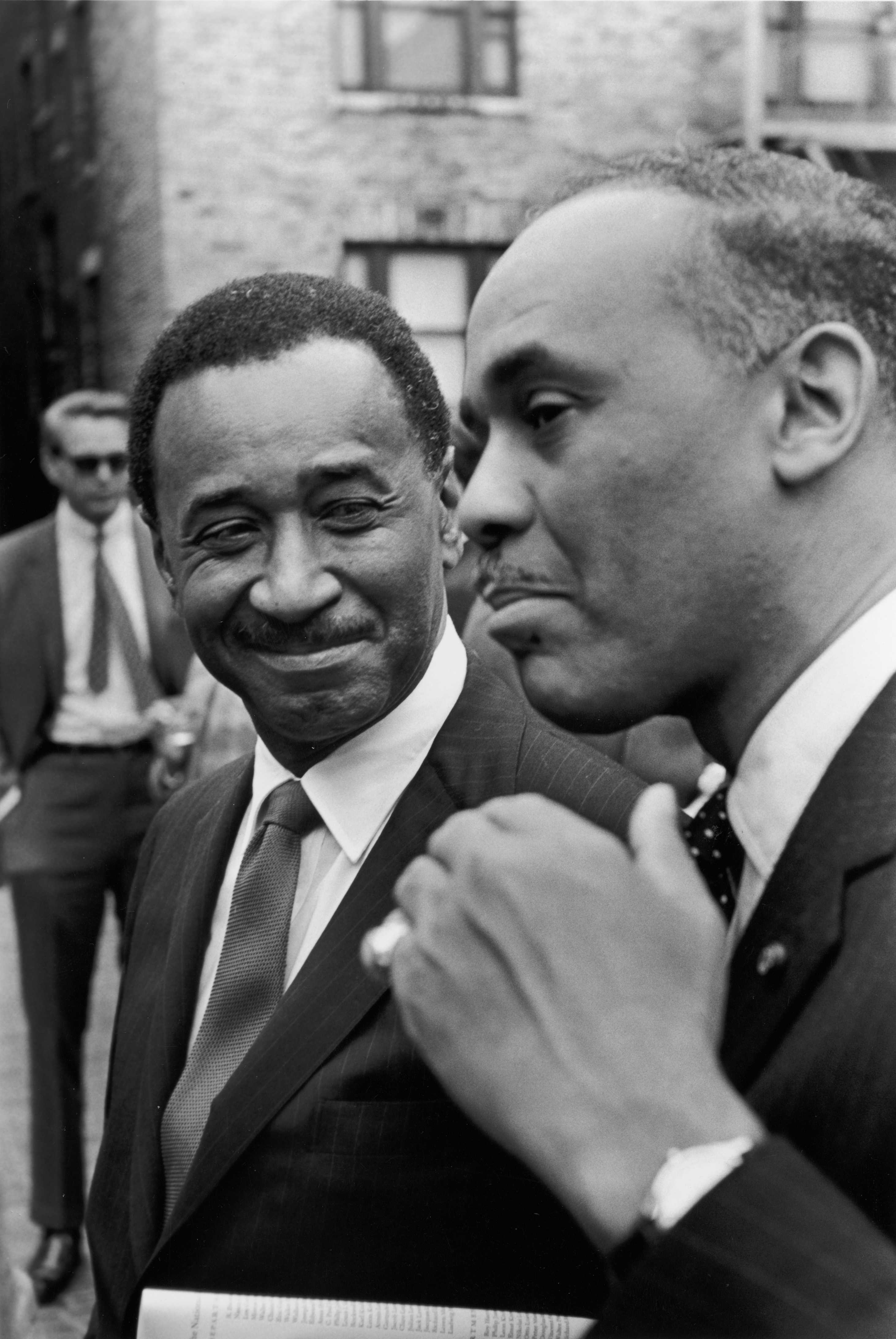 Two individuals in formal attire are engaged in conversation outdoors.