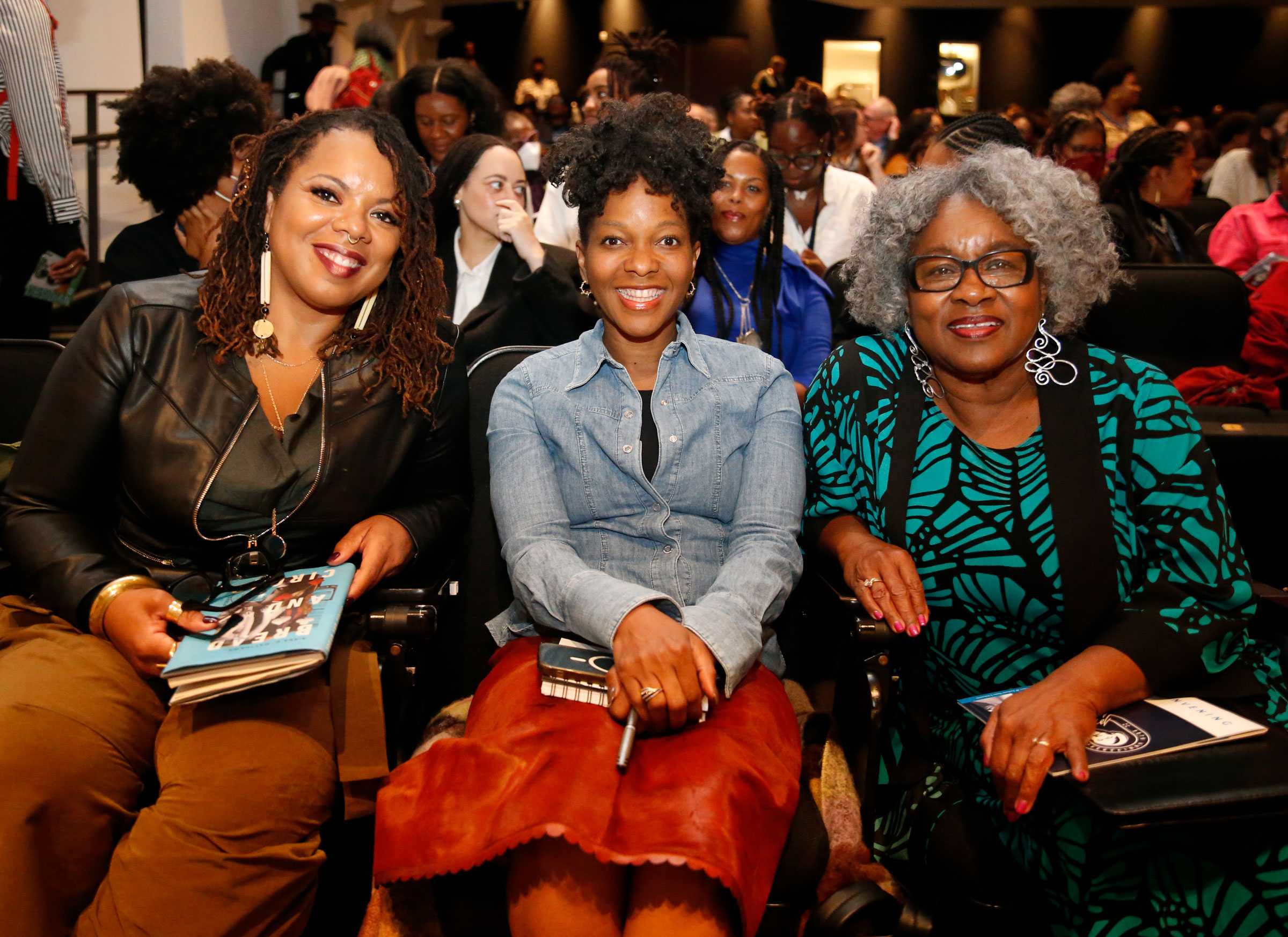 Three women seated in an audience, smiling,holding notebooks or books. People are gathered in the background.