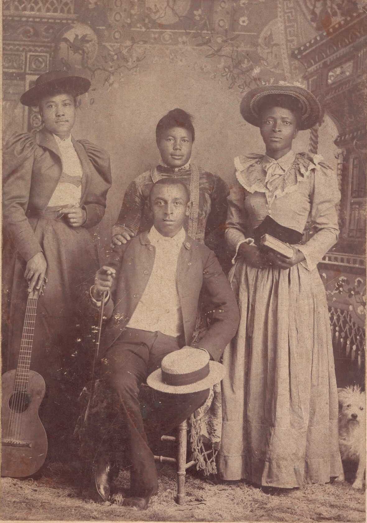 A cabinet card photograph depicting a sepia toned image of three women standing behind a seated man. The seated man is holding a cane with his right hand. His left hand is resting on his knee and holding onto a hat. The woman on the left side of the photograph is wearing a dark colored dress and jacket with a white shirt and dark colored hat. She is holding a guitar with her right hand. The woman in the middle has her right hand resting on the shoulder of the seated man. She is wearing a dark colored dress. The woman on the right side of the photograph is wearing a light colored, patterned dress and a light-colored hat. She is holding two books in her hand. Printed on the card beneath the photograph is “O. S. Goff” and “Fort Custer, Mont.” The back of the card is blank.