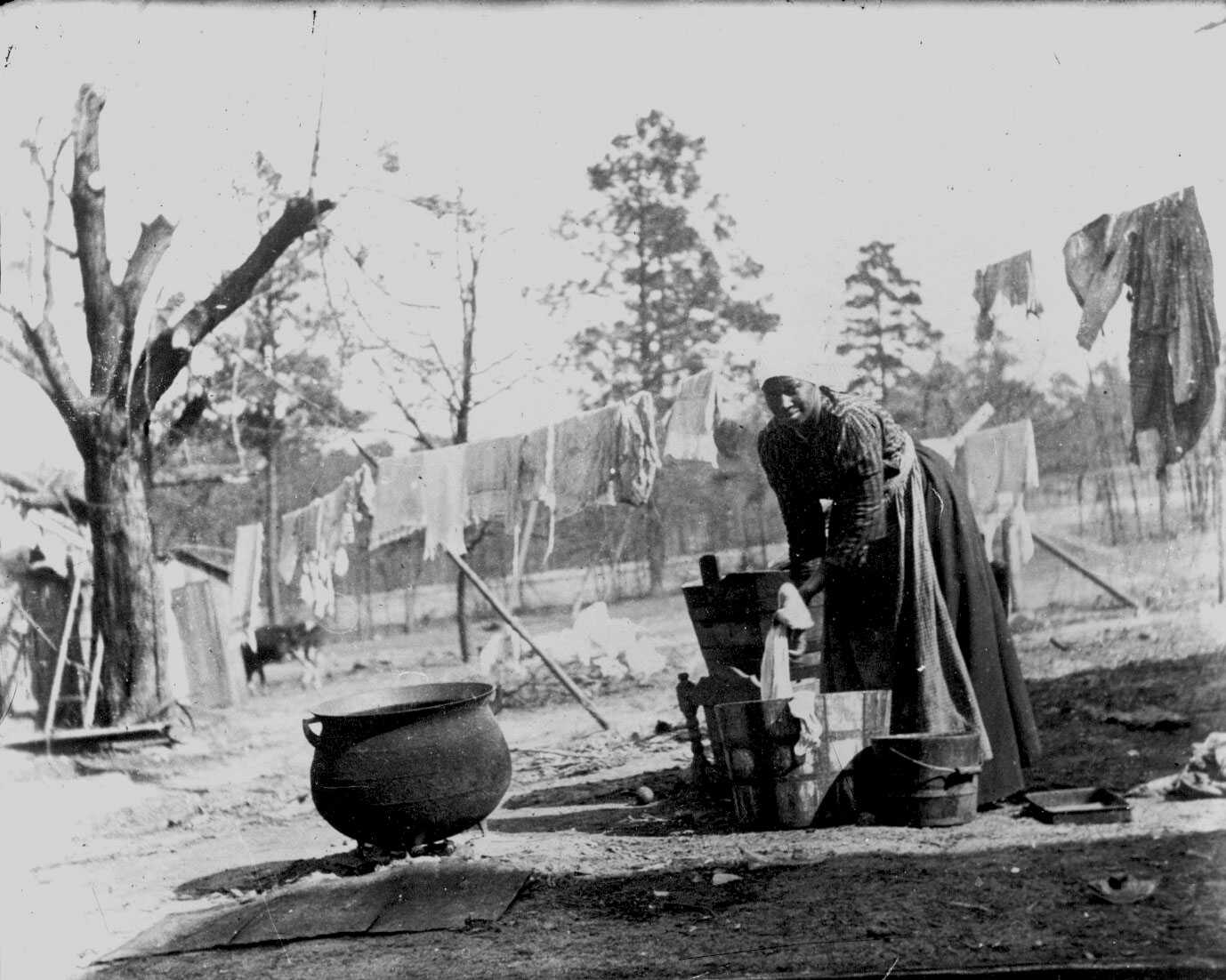 A photograph of an African American woman washing laundry outdoors in a yard. Clothes hand behind her on a line.