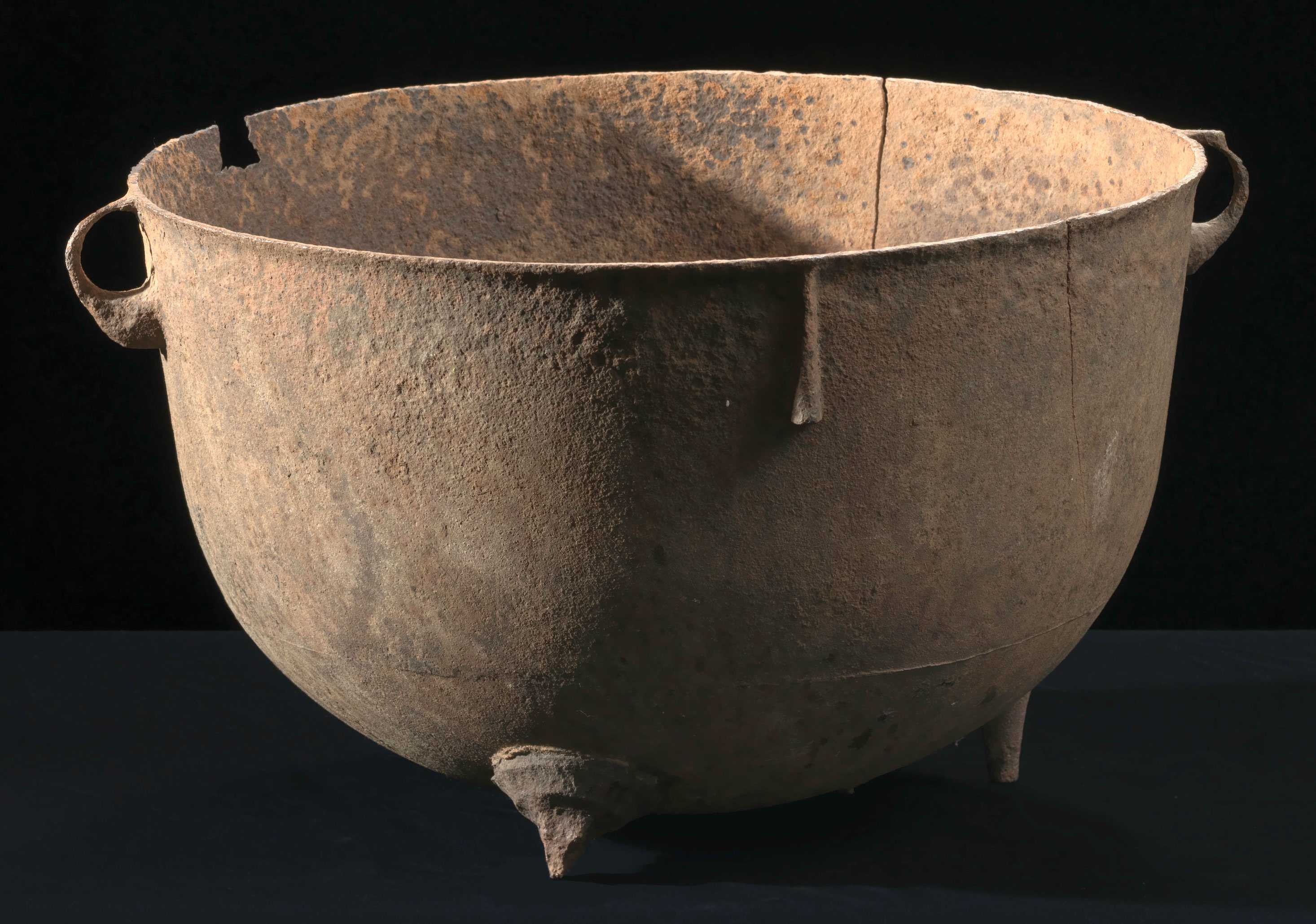 A large wash pot has small handles and small feet. It is worned from rust and dirt and cracked.