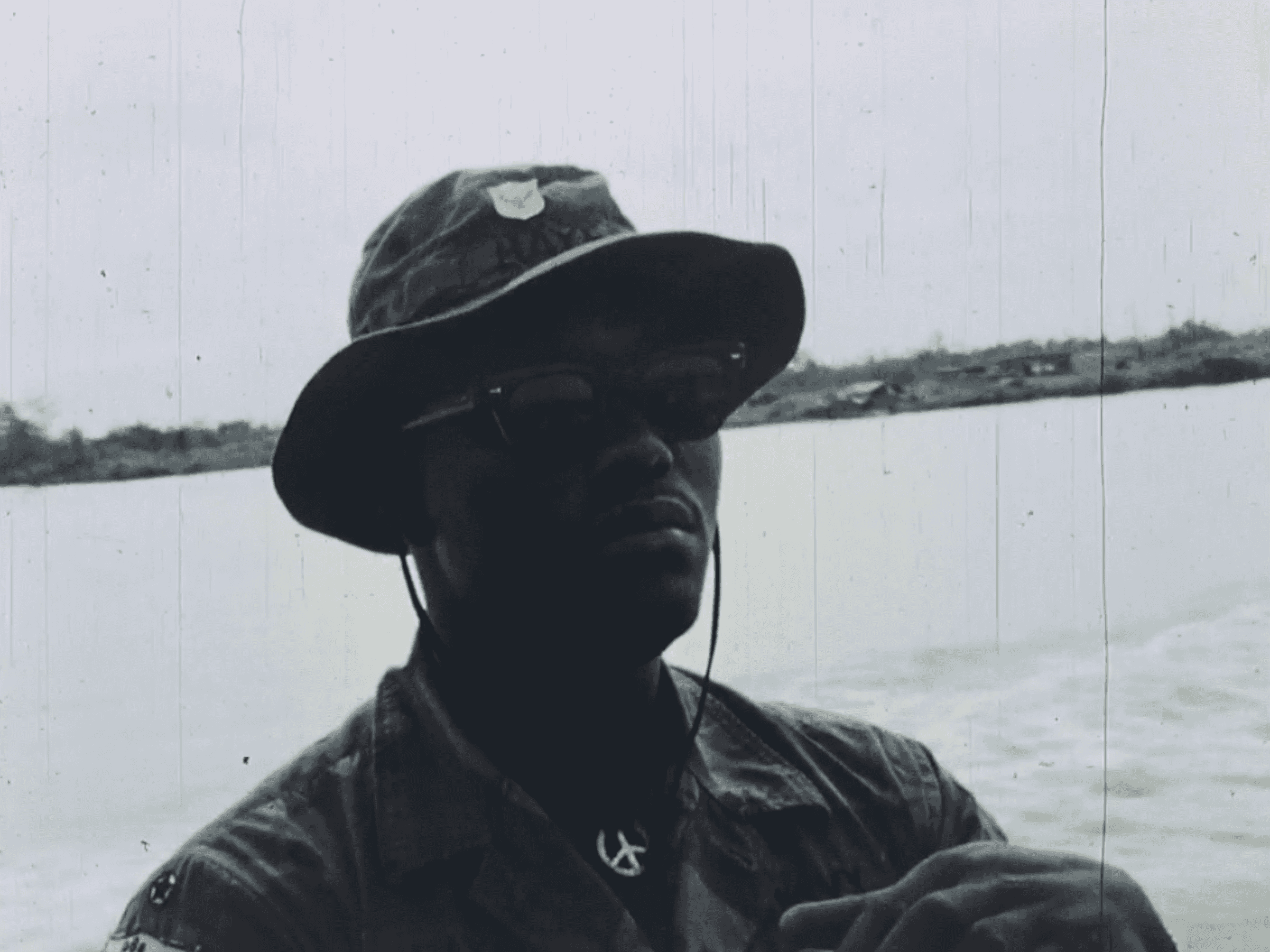 A soldier is on a boat in a bucket hat looking off over the water.
