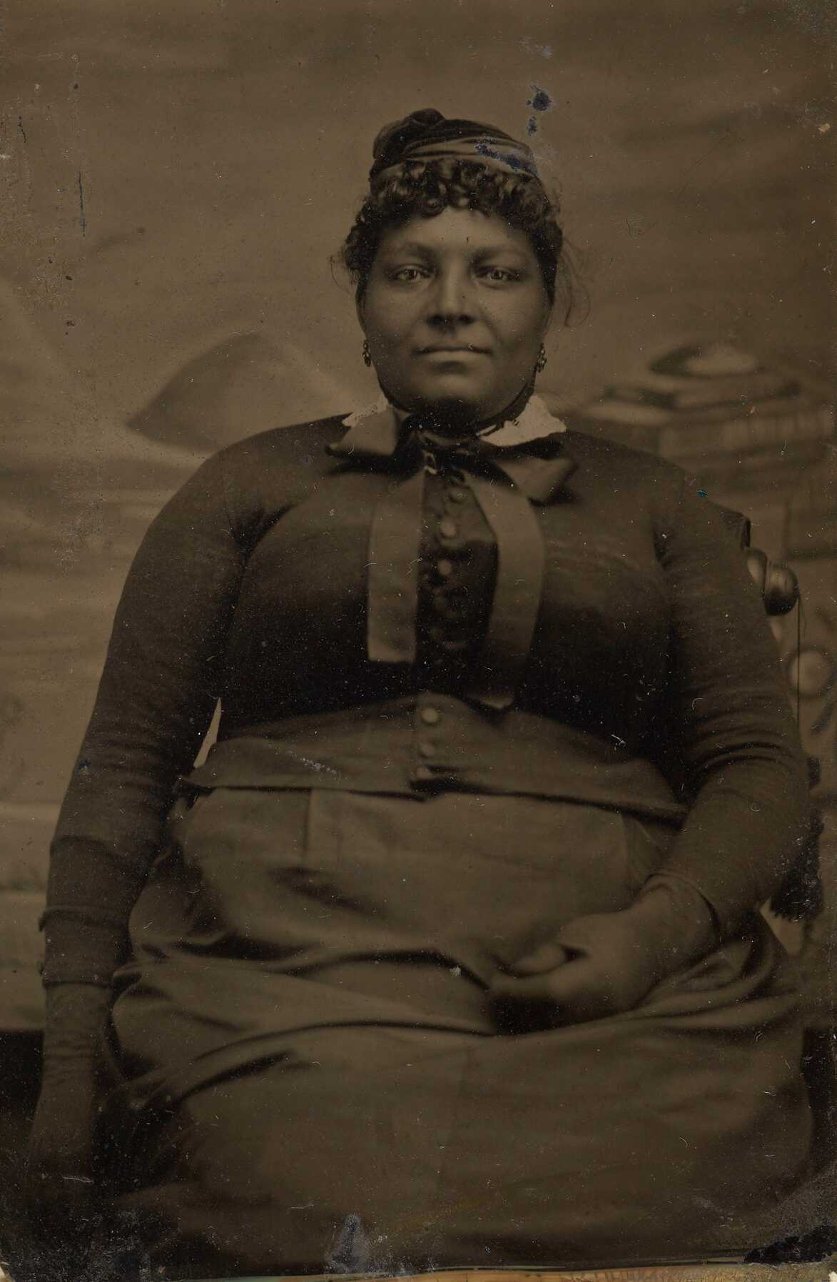 Tintype of Emmaline, a formerly enslaved woman. Emmaline is pictured seated in front of a studio backdrop. She wears a skirt, jacket and gloves of the same dark fabric. The jacket has a single row of small buttons up the center. She also wears a white collar with a dark ribbon, earrings, and a small hat. Her proper right arm is down by her side and her proper left hand is on her lap. She is smiling and is looking straight ahead at the camera. There are small losses to the tintype on the lefthand top and bottom corners.