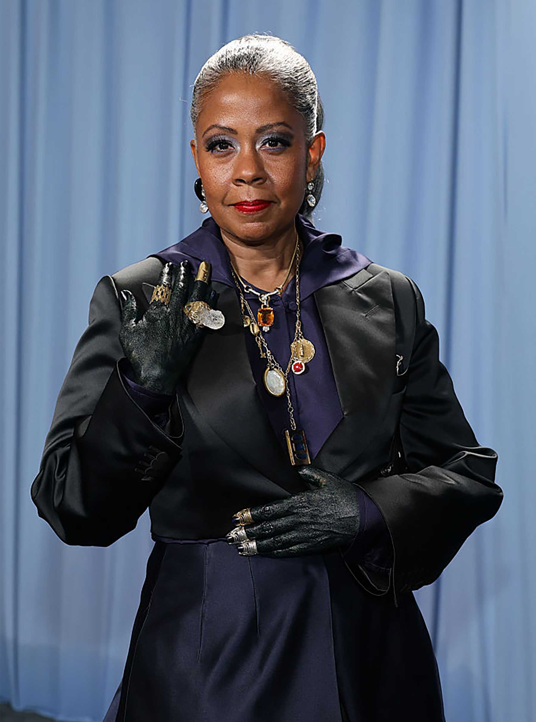 A woman in formal attire and jewelry poses against a curtain backdrop, with one gloved hand raised.