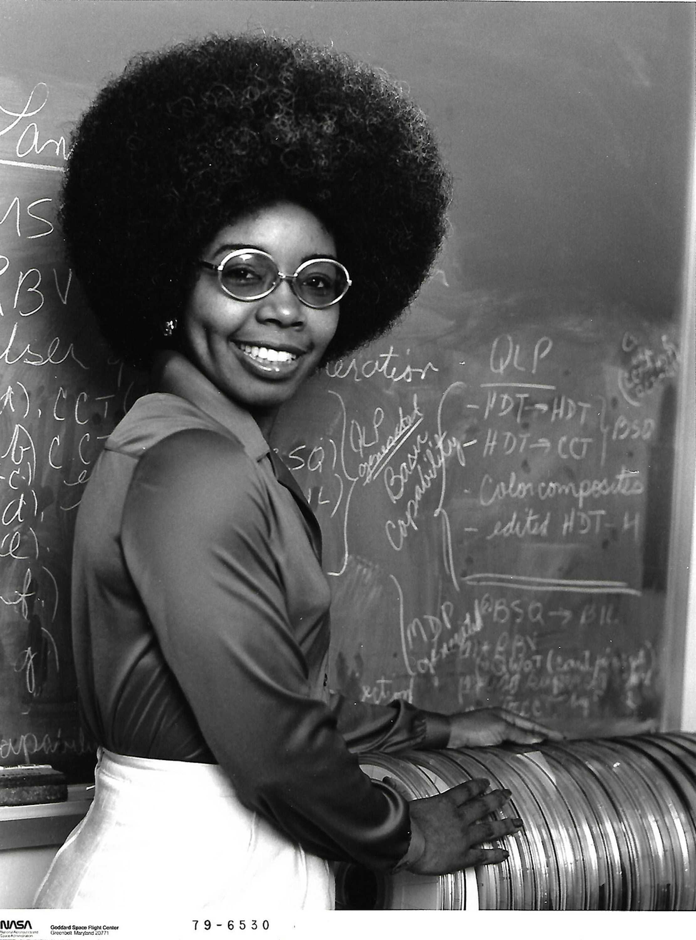 Valerie Thomas is smiling as she poses for a black and white photo in front of a chalkboard with writing on it.