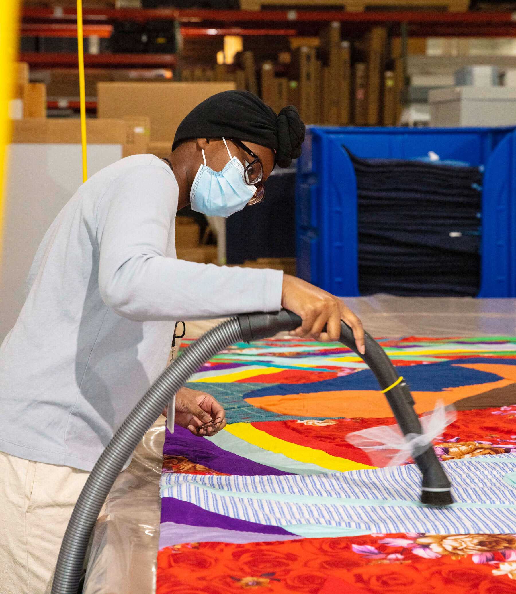 Masked person wearing glasses uses a hose vacuum to clean a colorful quilt on a worktable in a warehouse setting.