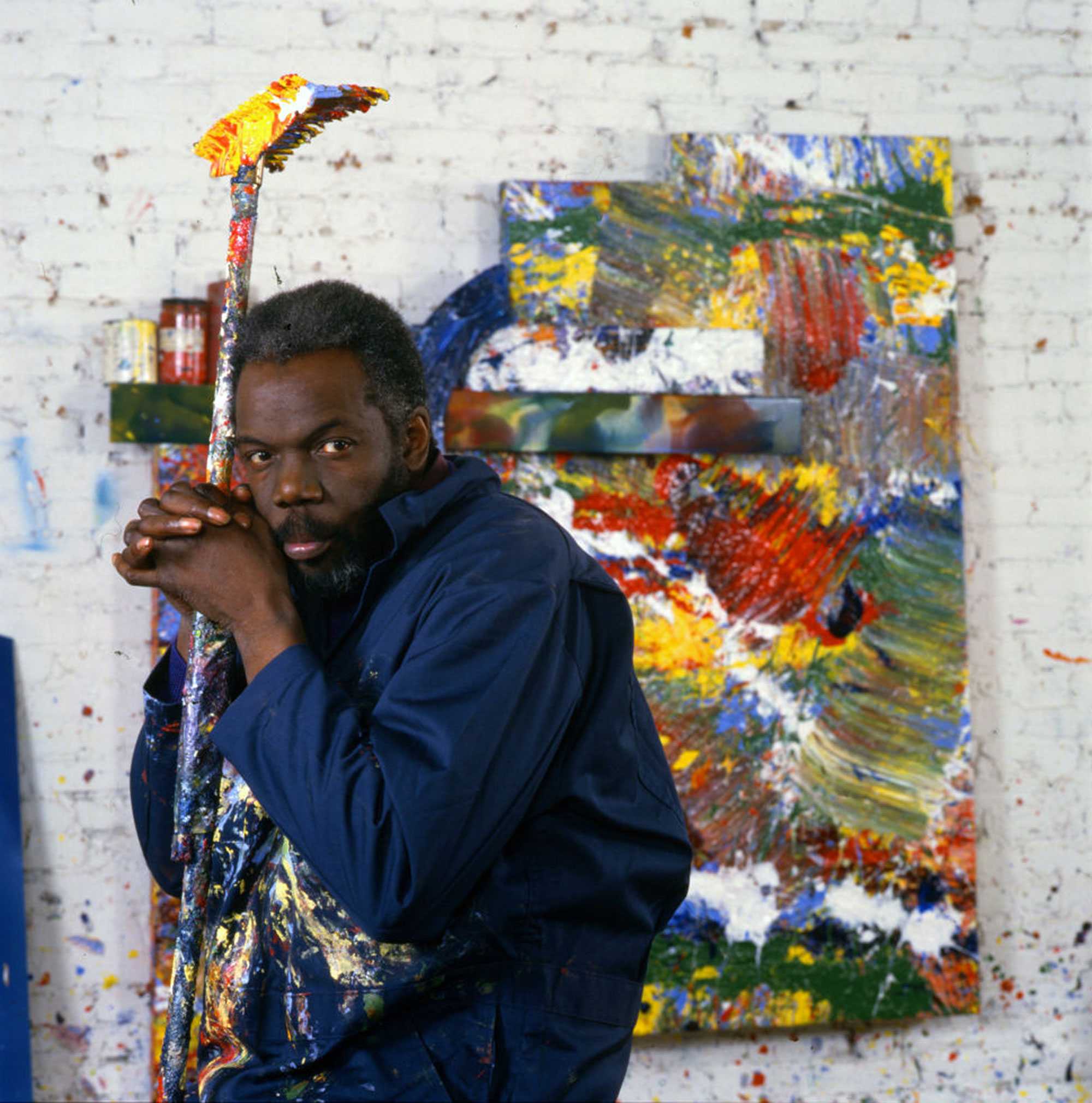 Artist in a studio, leaning on a paint-covered broom with a colorful abstract painting in the background.