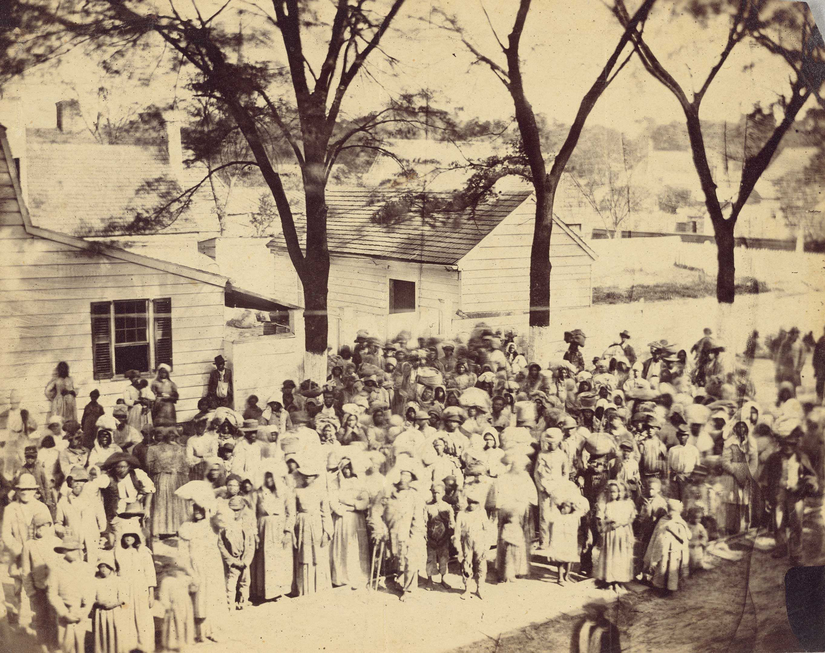 A worn black and white photograph that is slightly exposed of slaves standing in front of the cabins on J. J. Smith's cotton plantation near Beaufort, South Carolina.