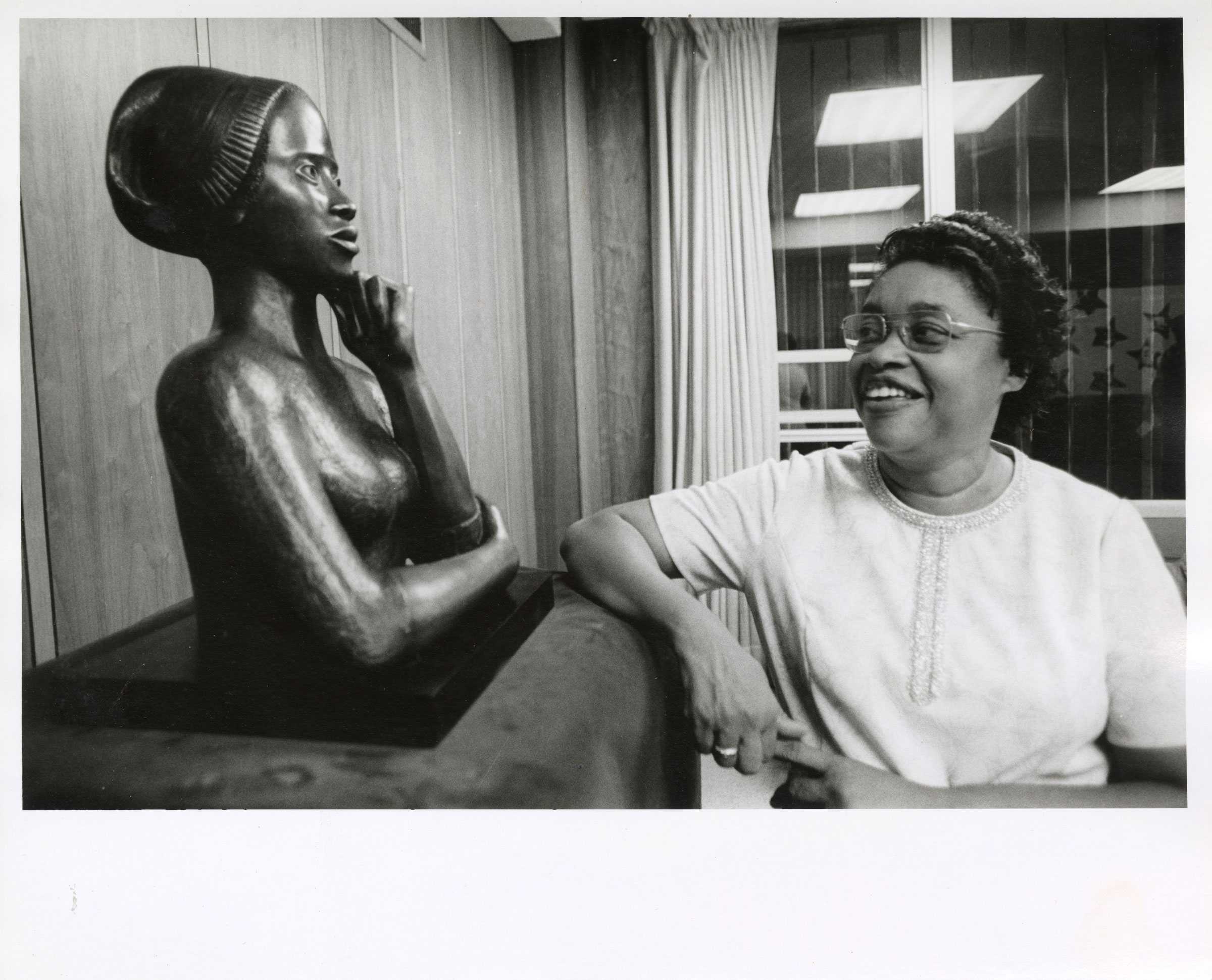 Black-and-white photo of a woman smiling at a sculpture of a bust in an indoor setting.