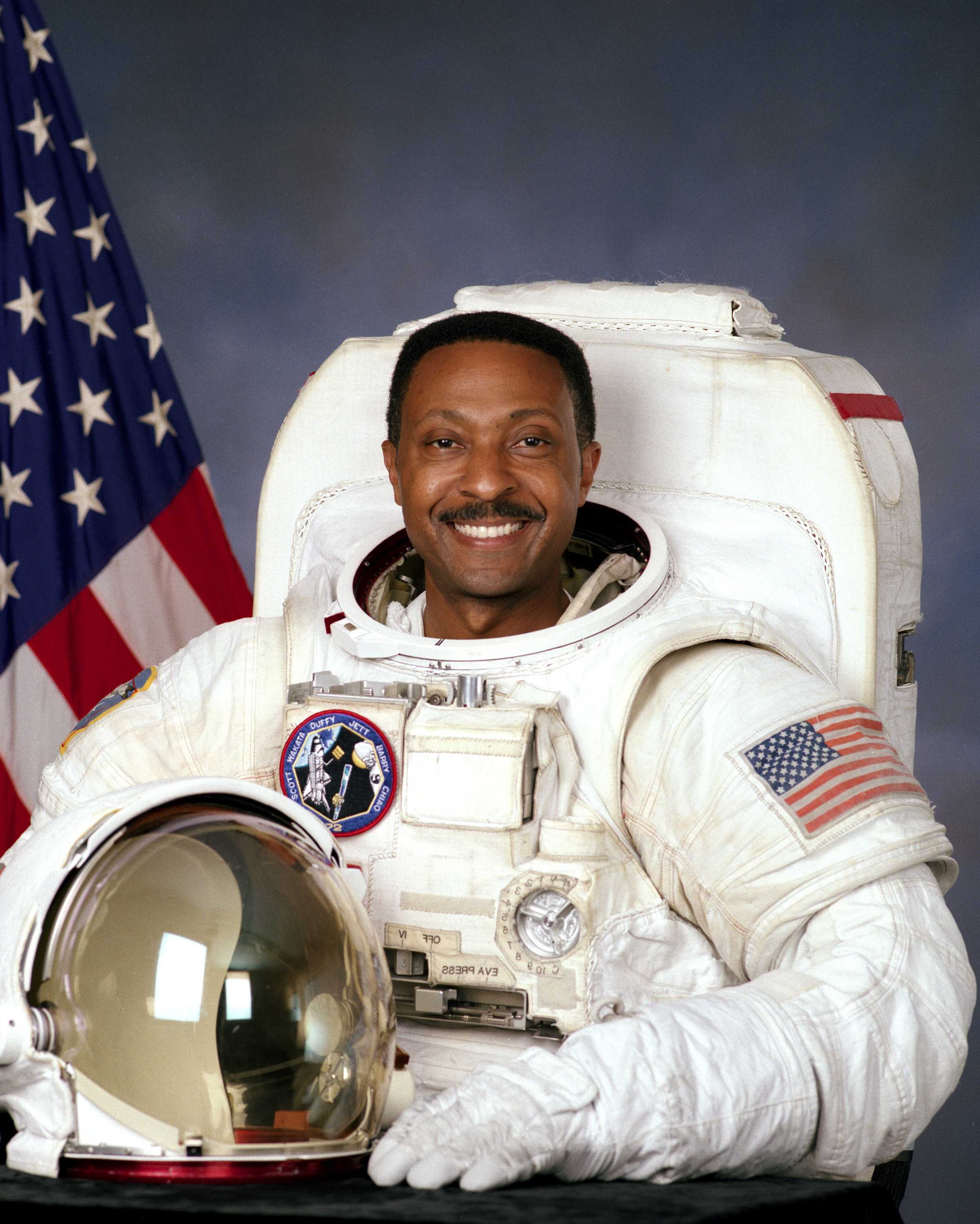 Winston Scott is wearing a white spacesuit adorned with NASA patches and smiling towards the camera.