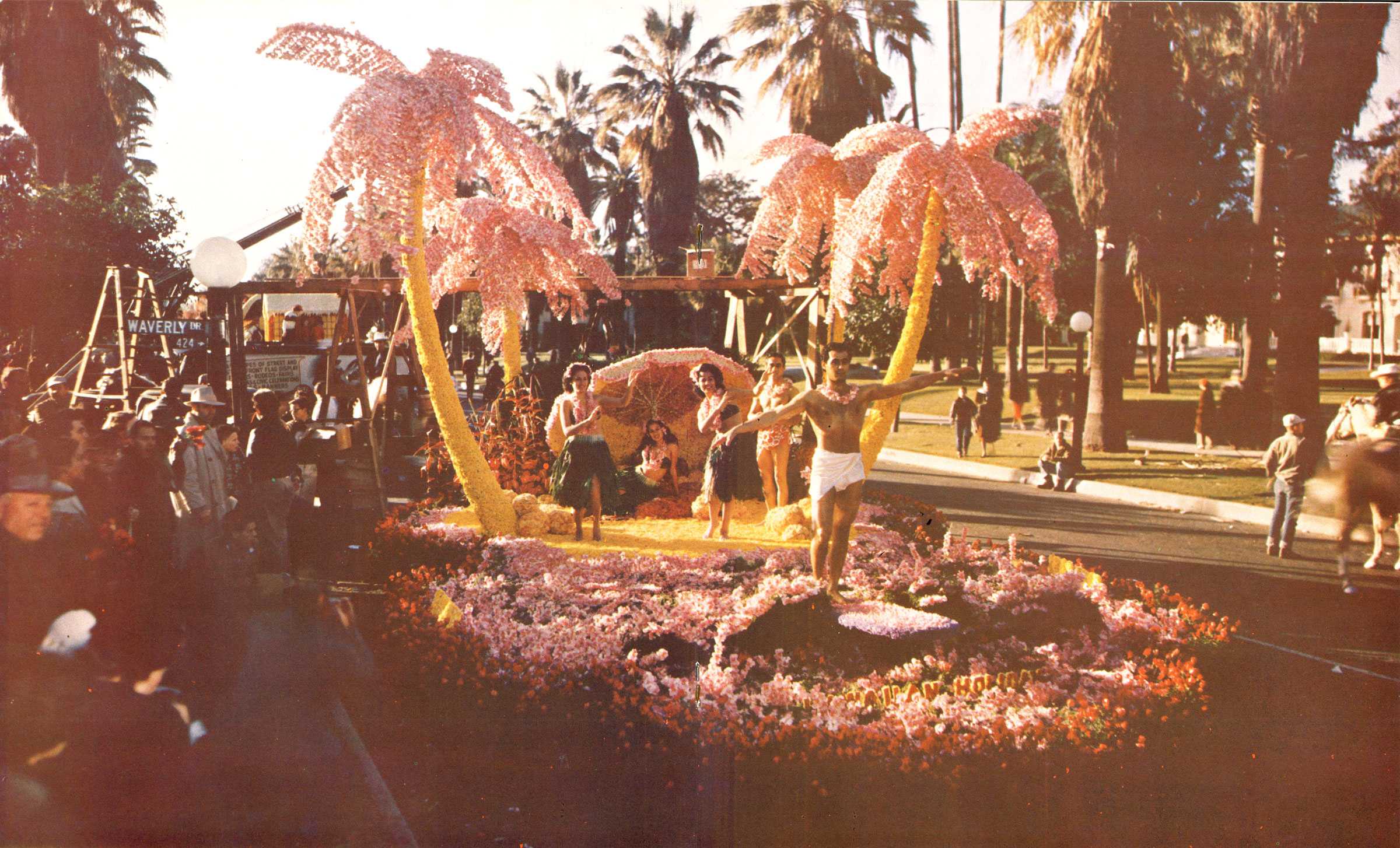 Parade float depicting a tropical scene with costumed people, two pink palm trees, and colorful flowers, observed by a crowd.