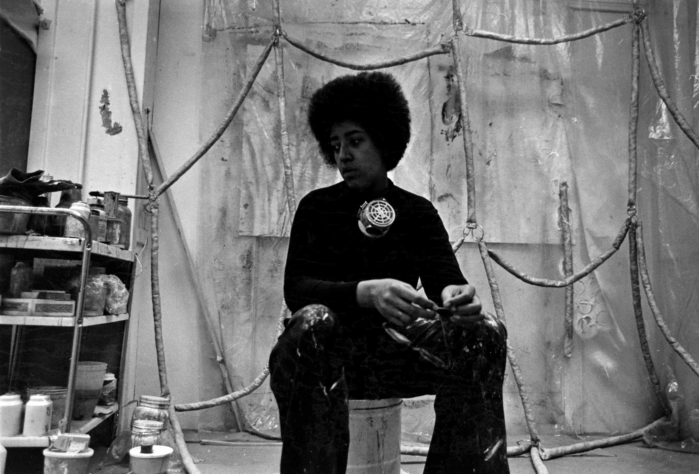 Black-and-white photo of a person sitting on a bucket in an art studio, with paint supplies and a protective mask around their neck.