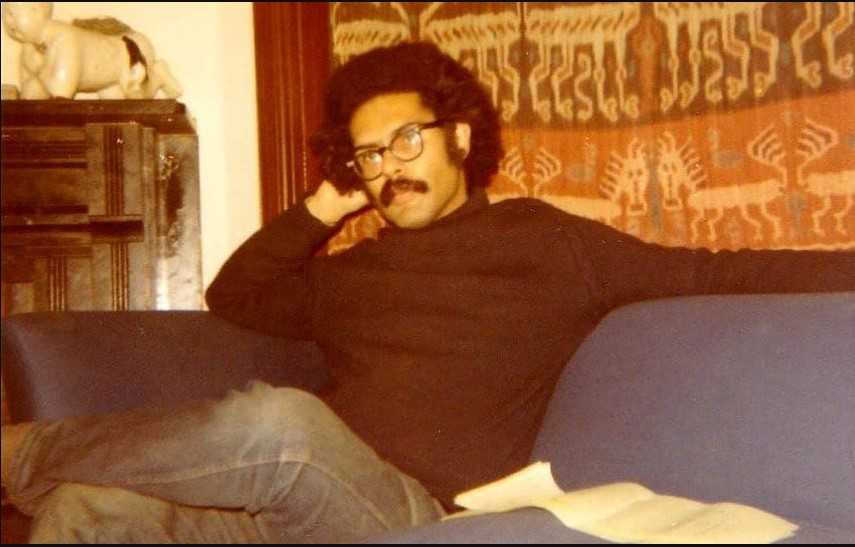 A colored photograph of Samuel Delany sitting on a couch wearing a black turtle neck and dark rimmed glasses.