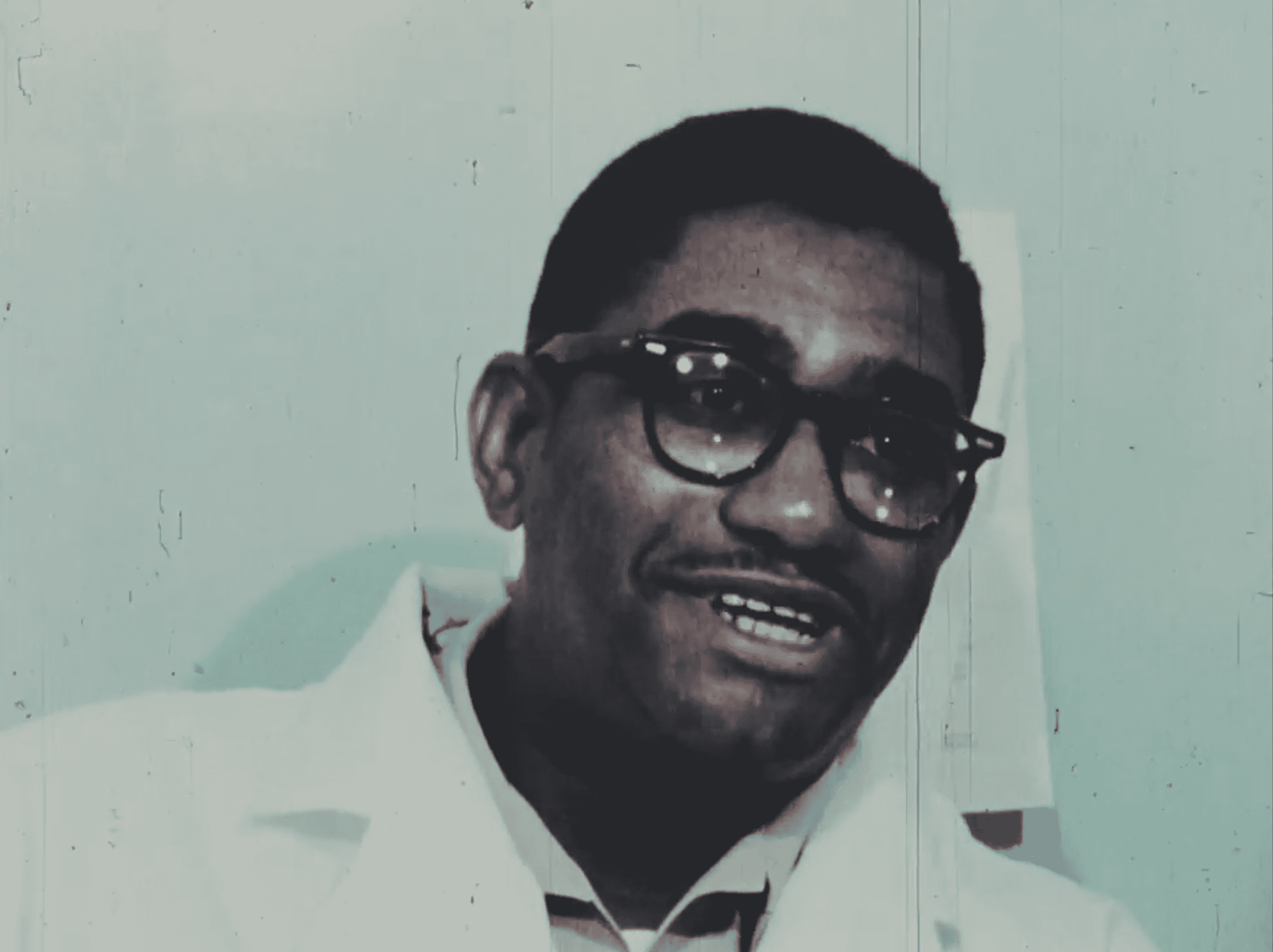 Man is talking while wearing a white collared shirt and thick rim black sunglasses.