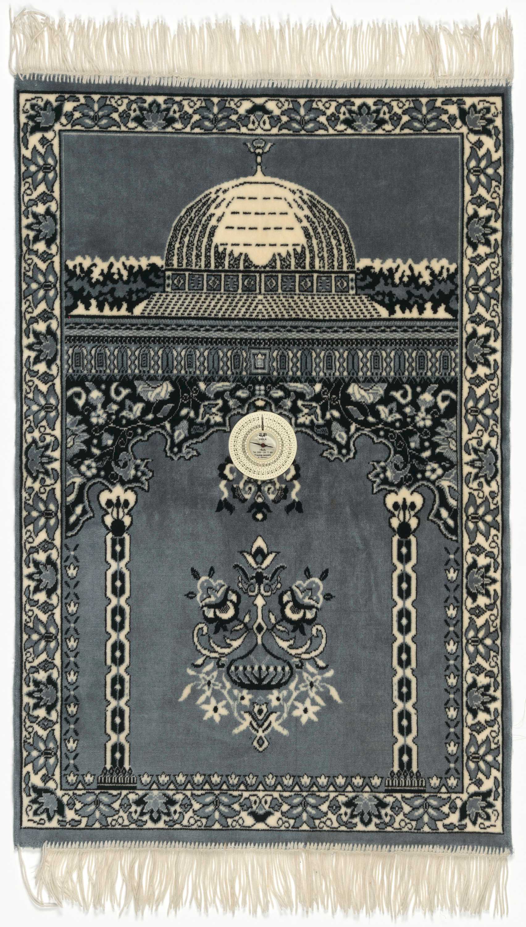 The prayer rug is a low-pile carpet with a primarily gray-blue background with black and white design details. The design featured in the top third of the rug is the image of an architectural dome with a small floral spire at the top. Below the dome is a geometric pattern running horizontally across the rug. Below the geometric pattern in the lower two-thirds of the rug is a stylized architectural interior with geometric columns on either side of the space. At the top of the columns is a stylized arch of floral and scalloped patterns. At the top of the arch, attached to the rug, is a white plastic compass. The compass is a Qibla directional compass. Below the compass needle, at the bottom, is the manufacturer information [HILAL / Tel. 069-28 77 69 / Praying carpets / W.-Germany]. Below the compass, in the center, is a floral motif. Surrounding the edges of the rug is a border with a floral pattern. At the top and bottom of the rug is white fringe.