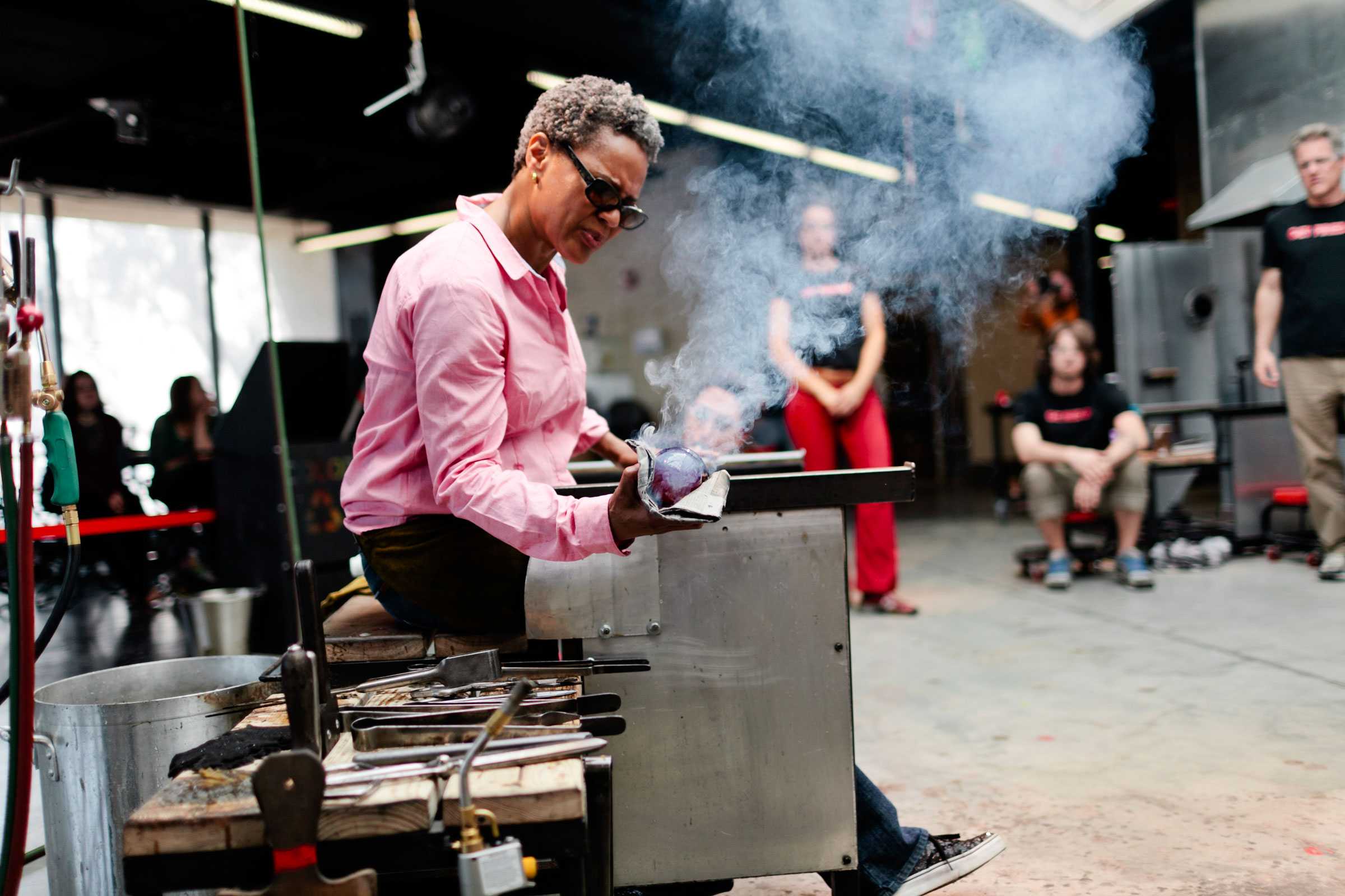 Person in a pink shirt working on a glassblowing project, with tools on a table and an audience watching in the background.