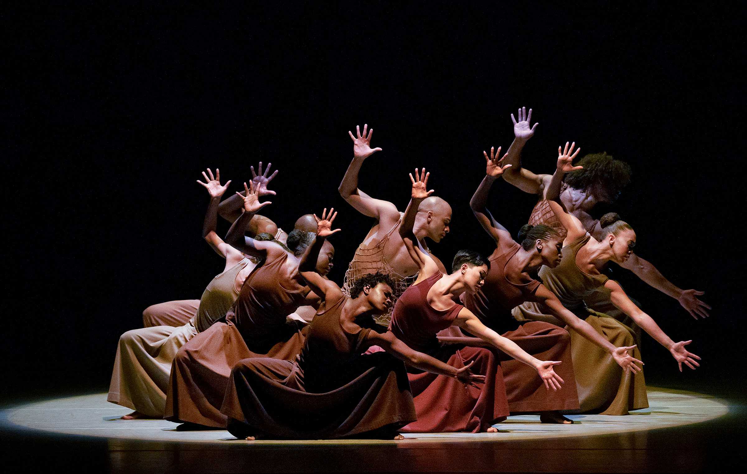Dancers in a group choreography on stage, wearing brown outfits, with hands raised dramatically against a dark background.