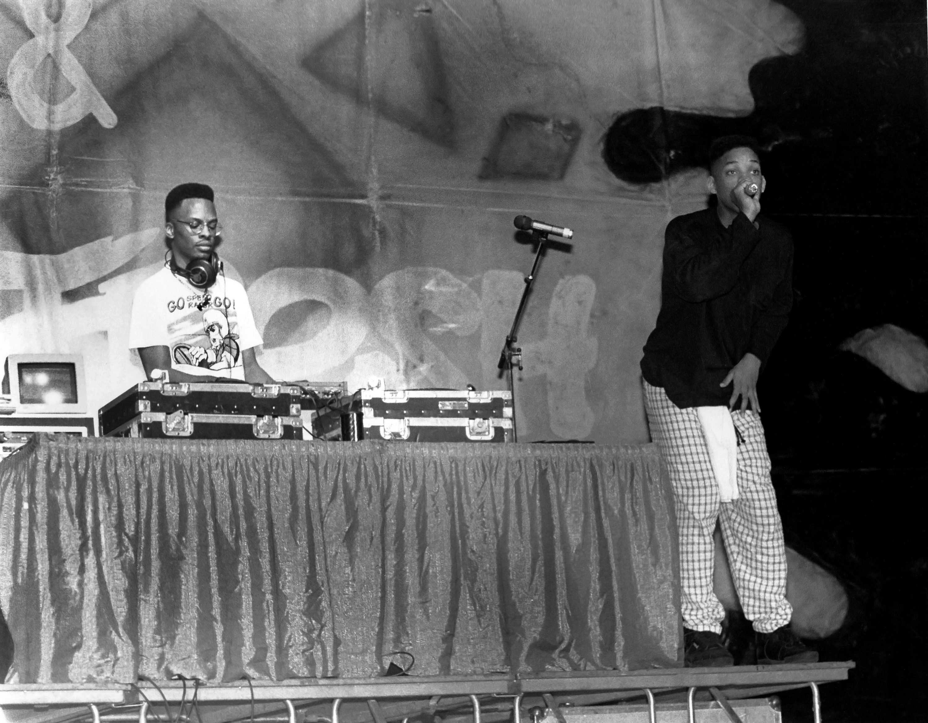 DJ Jazzy Jeff is behind a table, mixing a record, while Will Smith is rapping into a microphone.