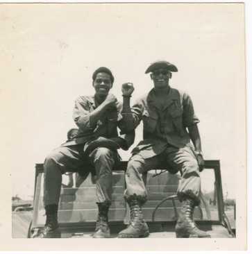 A black and white photograph of two soldiers, in military fatigues, perched on the edge of the windshield, boots resting on the jeep's hood.
