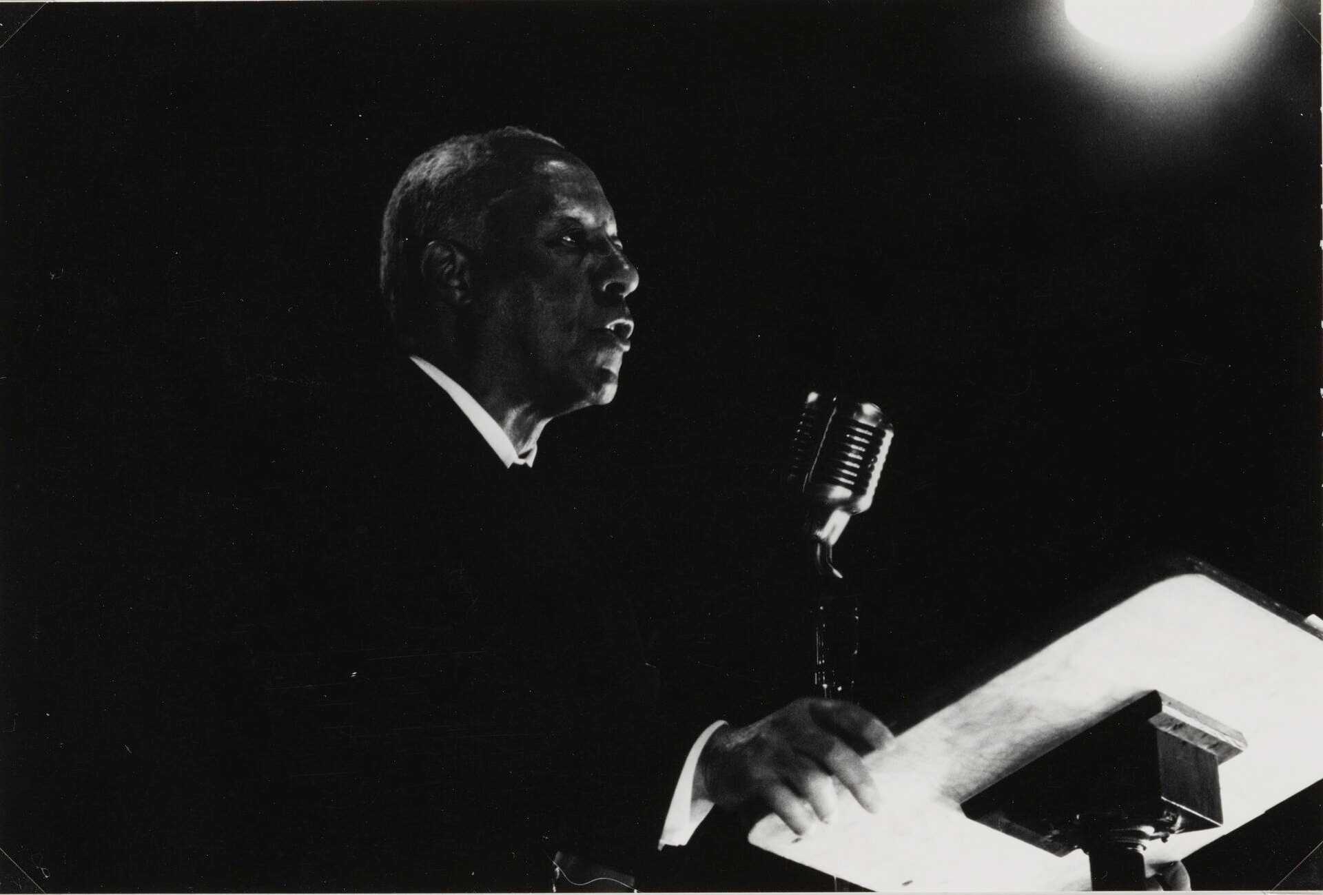 A silver gelatin print depicting a black-and-white image of A. Phillip Randolph giving a speech at an NAACP Convention in Albany, New York. The background is black with a single light illuminating from the upper right corner. Randolph is at the center, dressed in a black suit with a white dress shirt. He is standing in front of a microphone and podium. His proper right hand holds the edge of the podium and he is speaking into the microphone.

There are two inscriptions on the front of the print identifying the subject and the photographer's signature.  On the back of the print is a vertical line made with pencil.