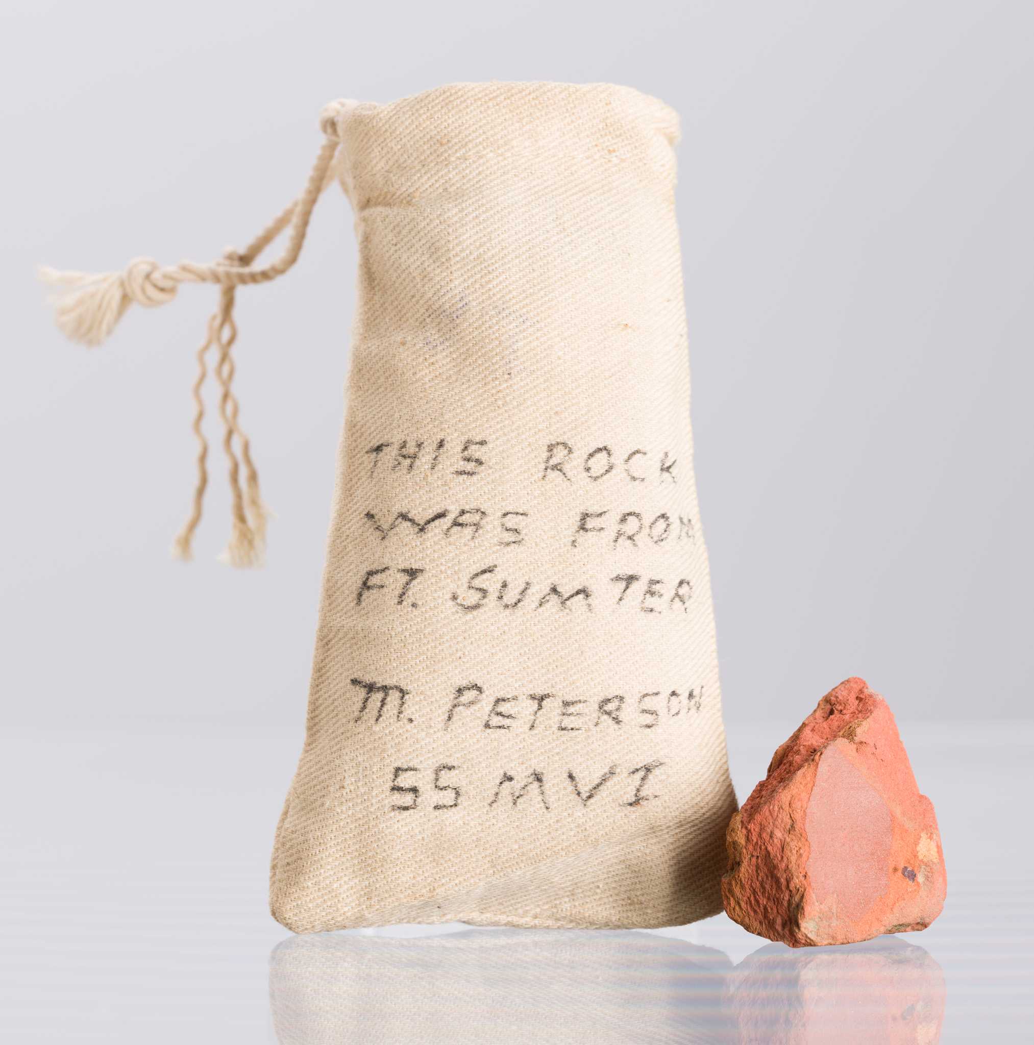 A cotton bag (2017.78.2a) containing a piece of brick (2017.78.2b) from Fort Sumter owned by Marquis Peterson. The bag is brownish white and has a braided drawstring at top. Hand written in black ink on the front of the bag is “THIS ROCK / WAS FROM / FT. SUMTER / M. PETERSON / 55 MVI.” The brick is reddish brown and is flat on the top and bottom. The four sides are irregular in shape and rough. The brick piece has several cracks throughout.