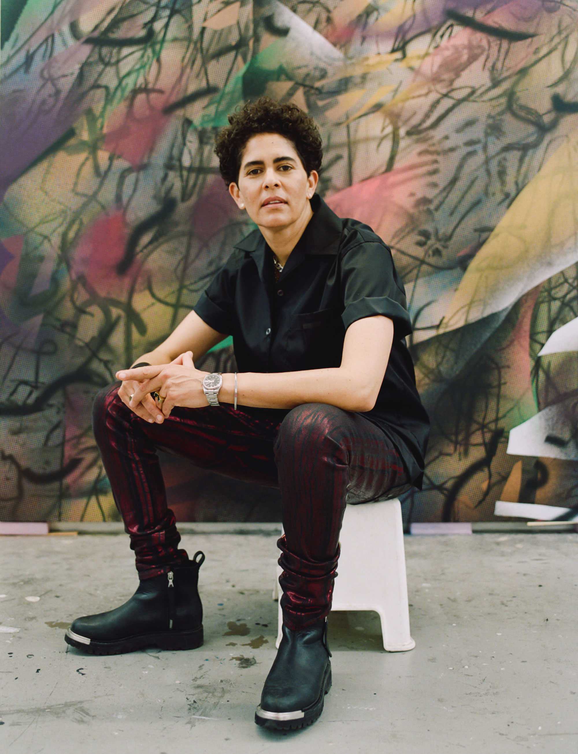 Person sitting on a stool in front of a colorful abstract mural, wearing a black shirt and patterned pants.