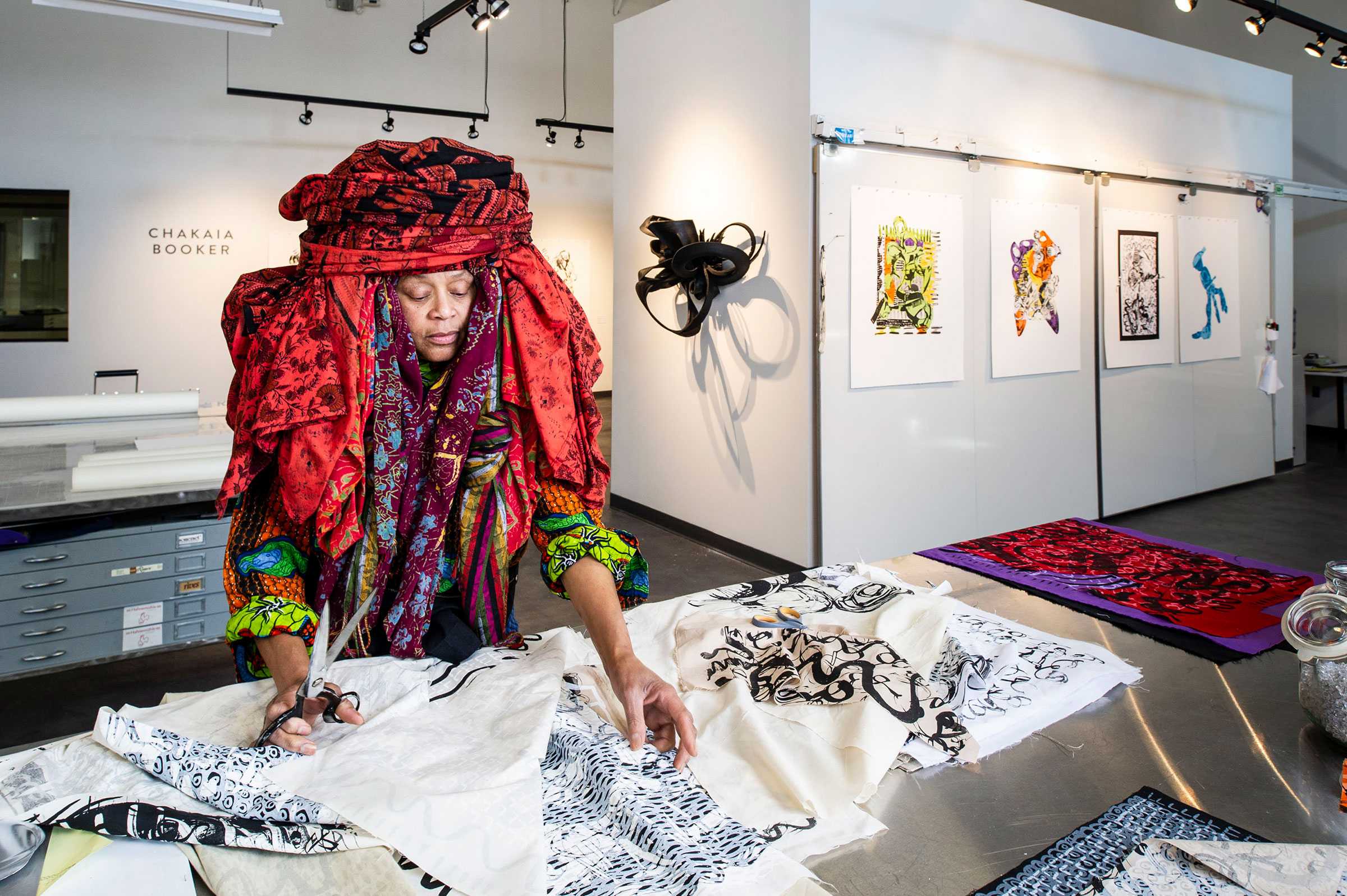 Artist in a studio, wearing an elaborate headpiece, cuts fabric with patterns. Art pieces are displayed on the walls in the background.