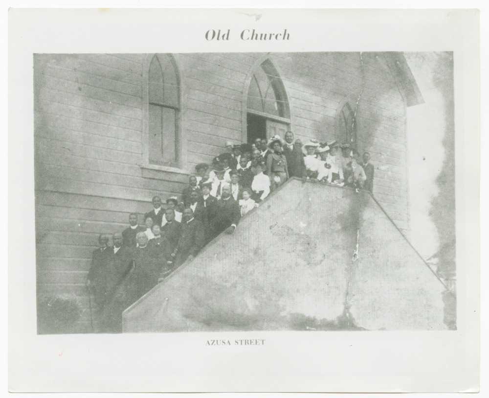 Black and white image of a copy of an image of people standing outside the steps of a church. The church exterior is wood paneling and features two (2) windows with gothic arches in the front. Between the two (2) windows, centered, is the doorway, also with a window above with a gothic arch. The arches are all divided by curved leading. The steps in the front of the building are obscured by a wall. The people stand on the steps between the exterior of the church and the exterior wall of the steps. The crowd of people are both men and women in a variety of clothes. The image is bordered in white with text centered at the top and bottom. At the top [Old Church]. Along the bottom [AZUSA STREET]. On the reverse in the lower left corner is a stamp with text and blanks to fill in. Only the last line is filled with a penned note [M. Matthews]. The stamp is four (4) lines [COLLECTION OF MIRIAM MATTHEWS/PHOTOGRAPHER/SOURCE/PLEASE CREDIT]. Along the bottom in the lower left is a penned note [AZUSA STREET CHURCH (FIRST A.M.E. CHURCH)]. In the upper right corner is the number [2] circled.