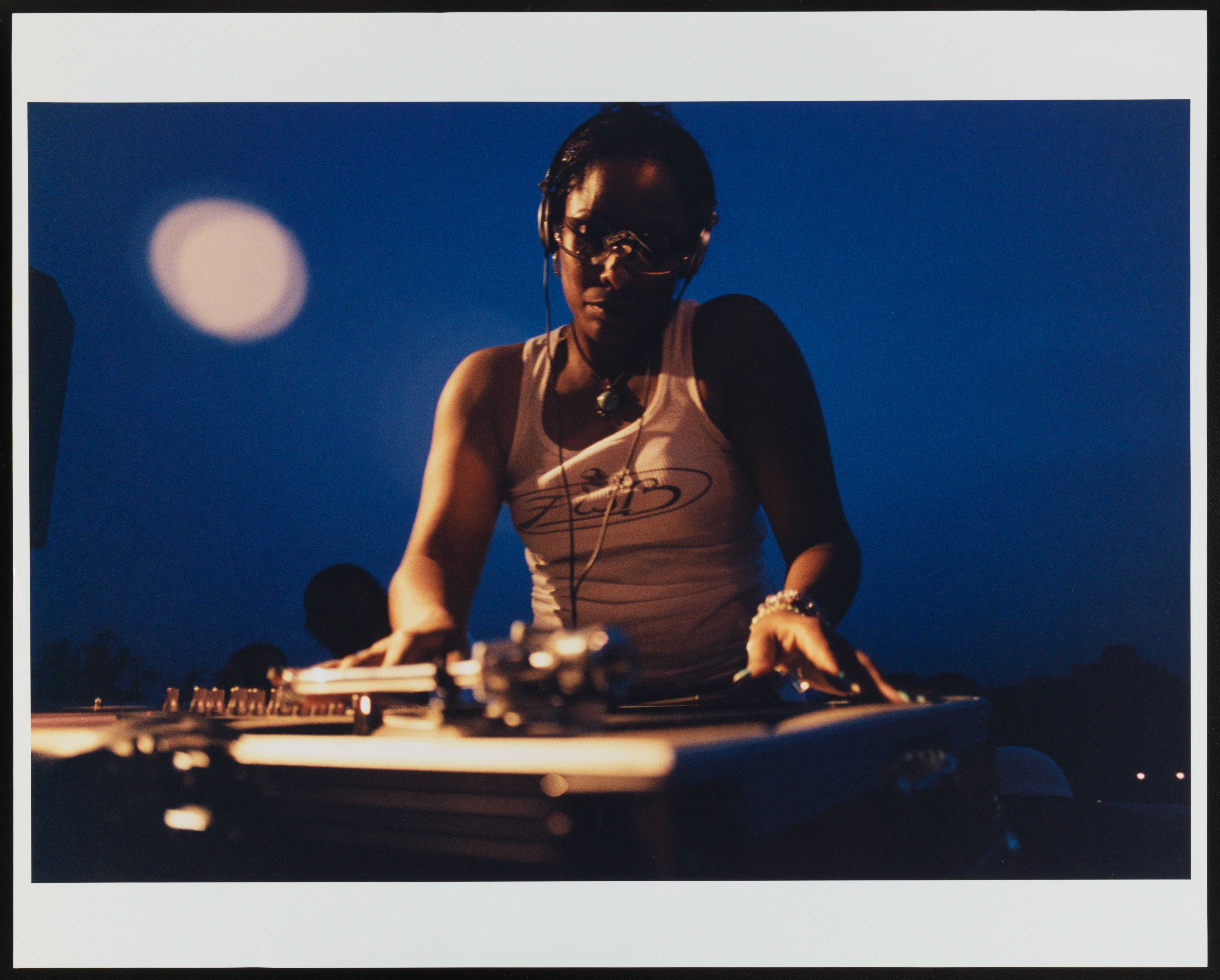 African American woman behind a turntable deck.  She is wearing a light colored tank top, headphone, glasses and a necklace.  A round of light is out of focus in the top left background.