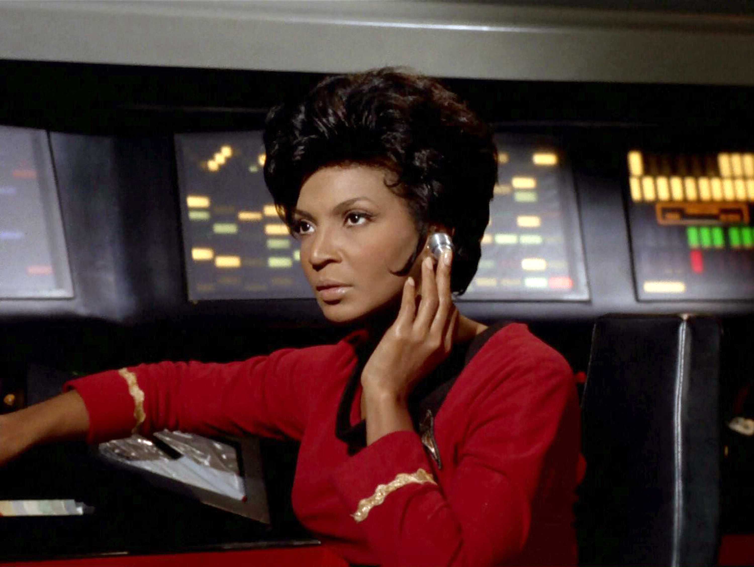 Lt. Uhura looking at off screen, holding an ear piece, in a read dress on a starship.