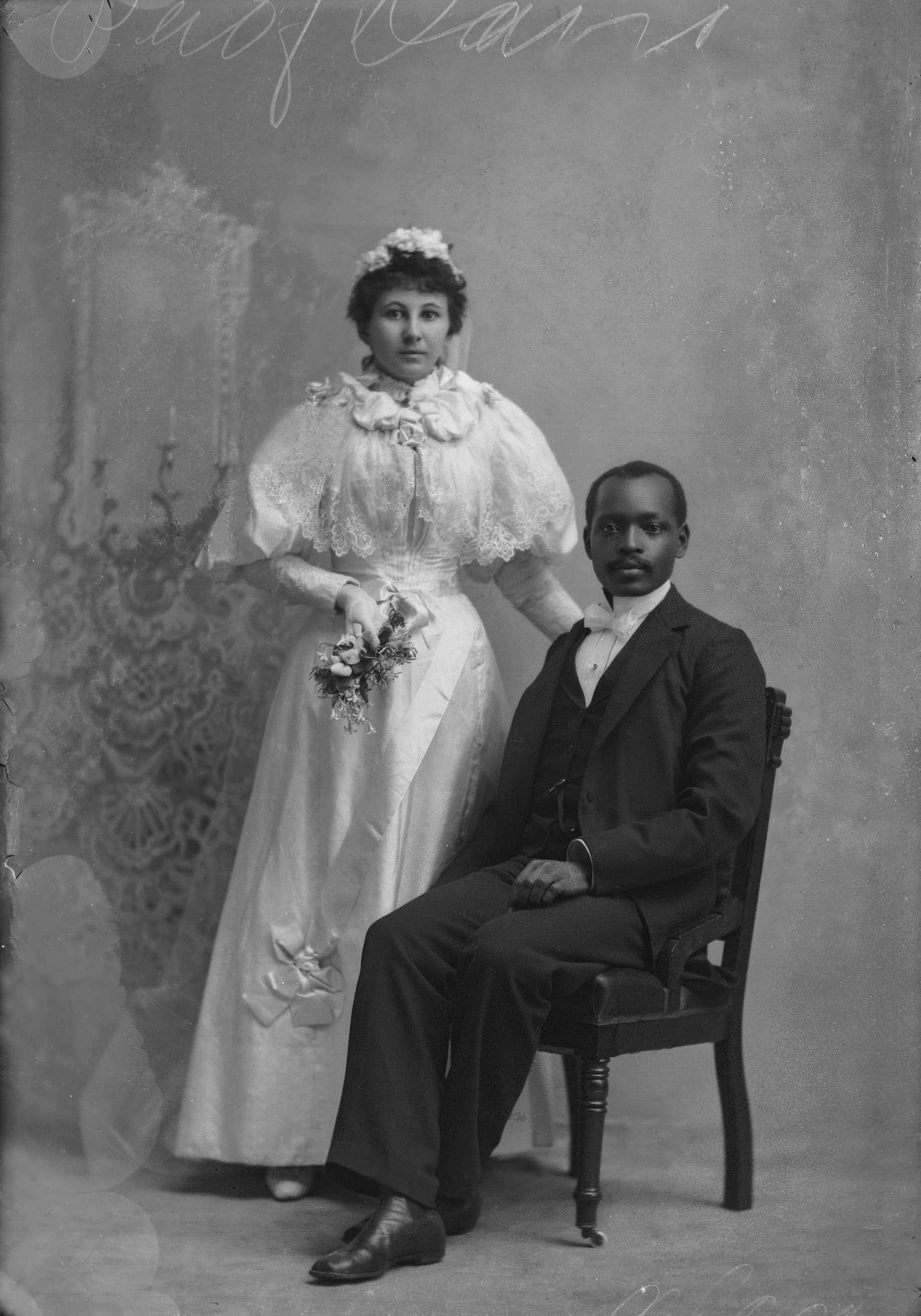 A worn black and white studio portrait of a bride and her groom. He is sitting in a chair when she stands.