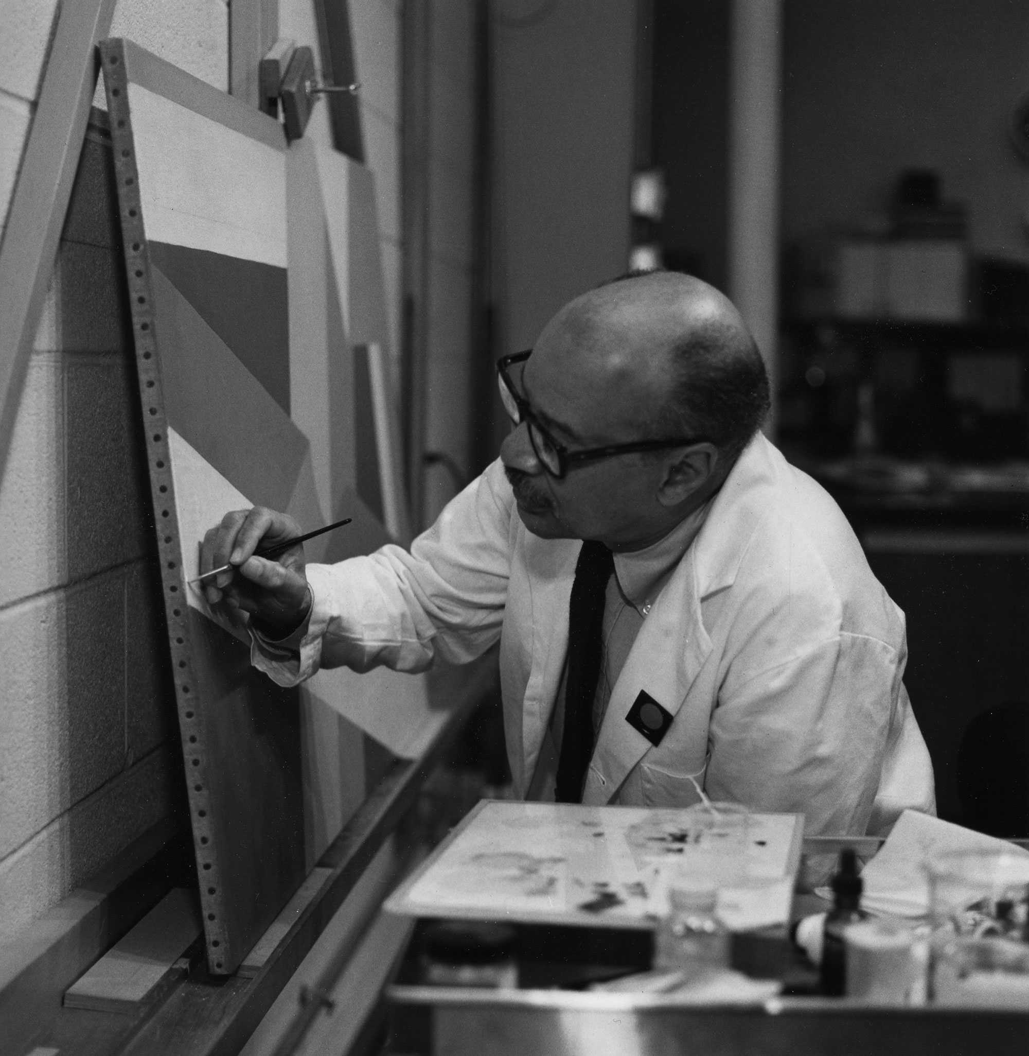 Man in a lab coat painting a geometric pattern on a canvas in a studio setting.