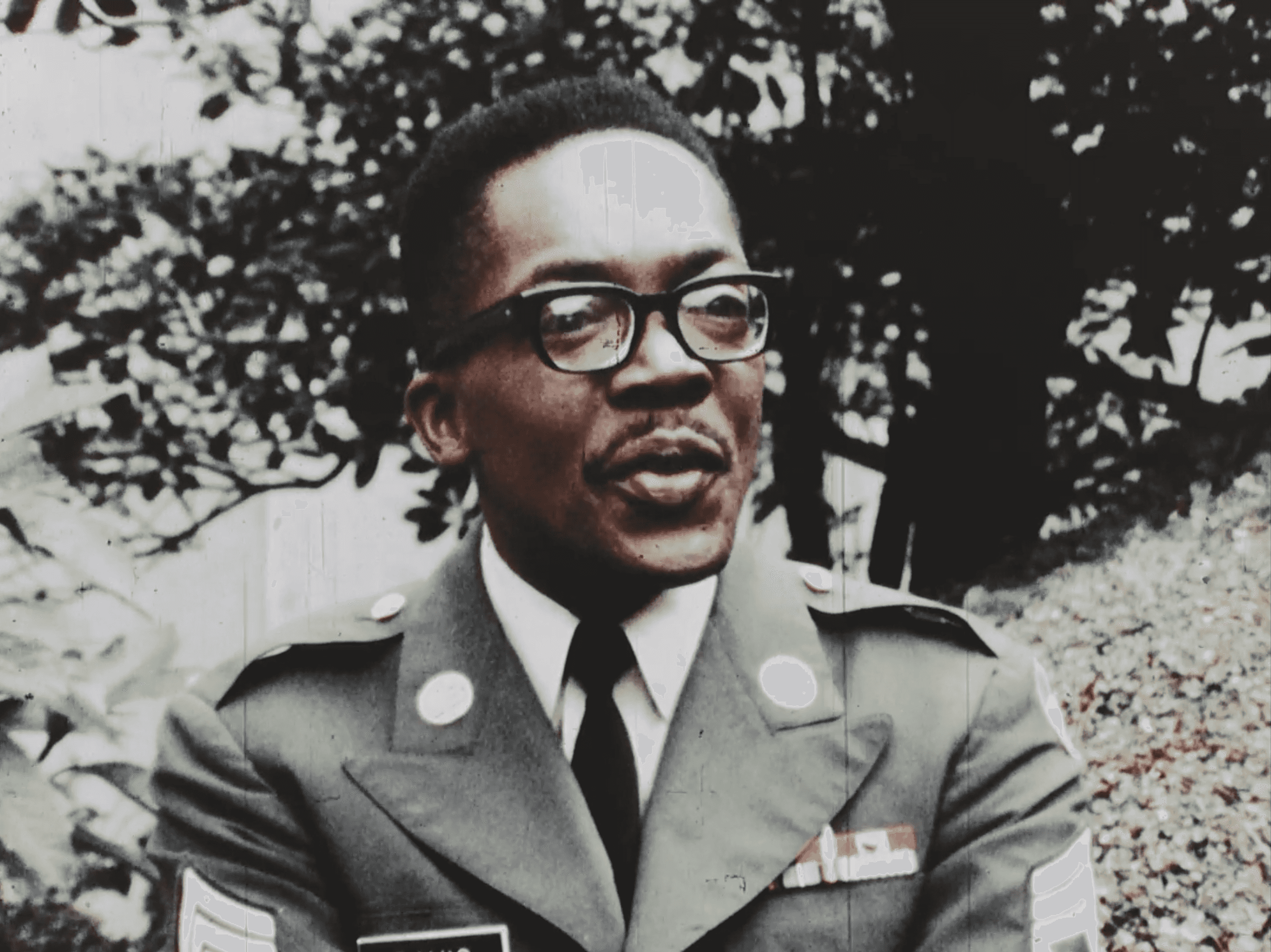 A man dressed in full military uniform is outside with trees and other greenery around him.